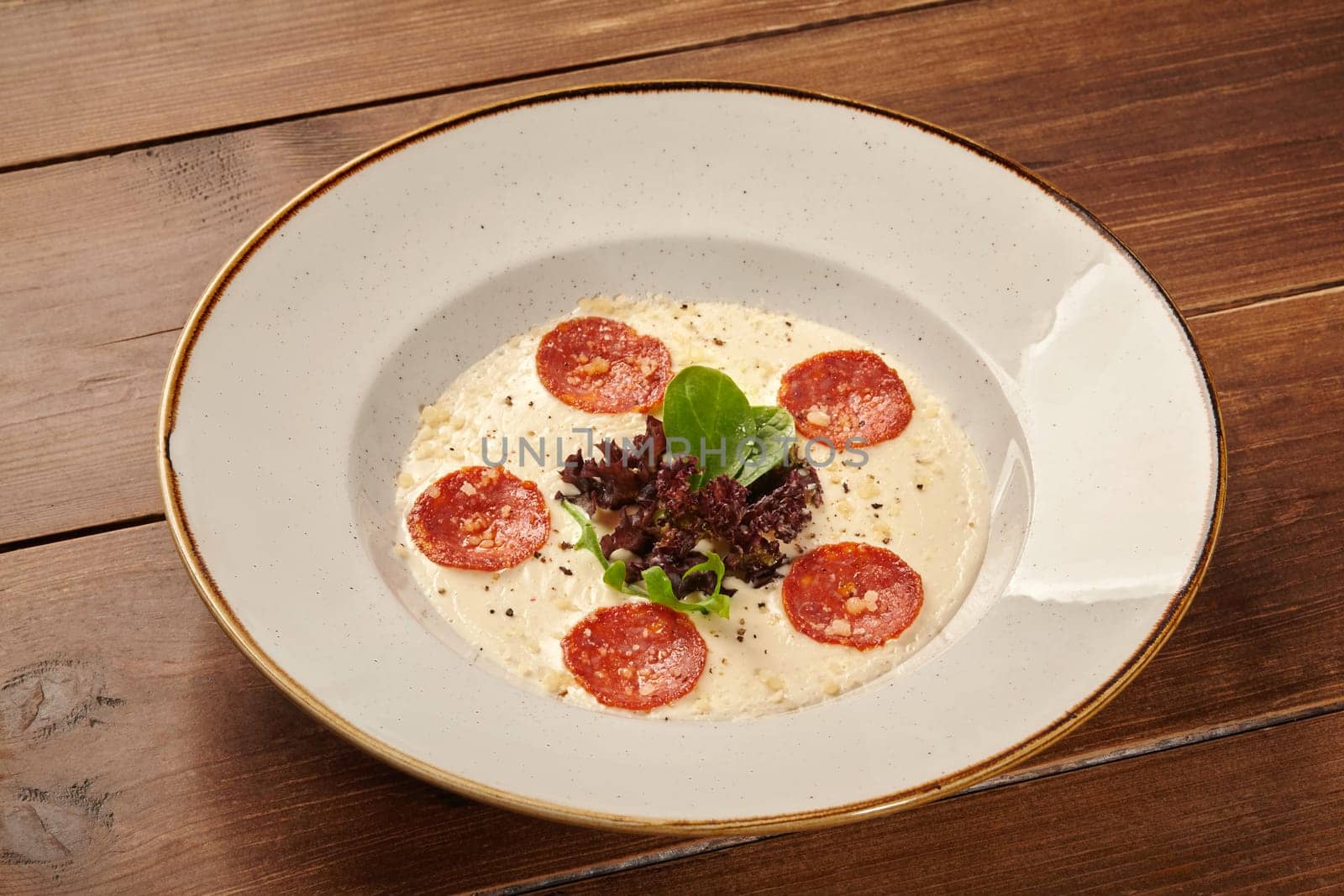 Creamy polenta topped with sliced pepperoni, parmesan cheese, and fresh herbs, served in a modern bowl on a rustic wooden table