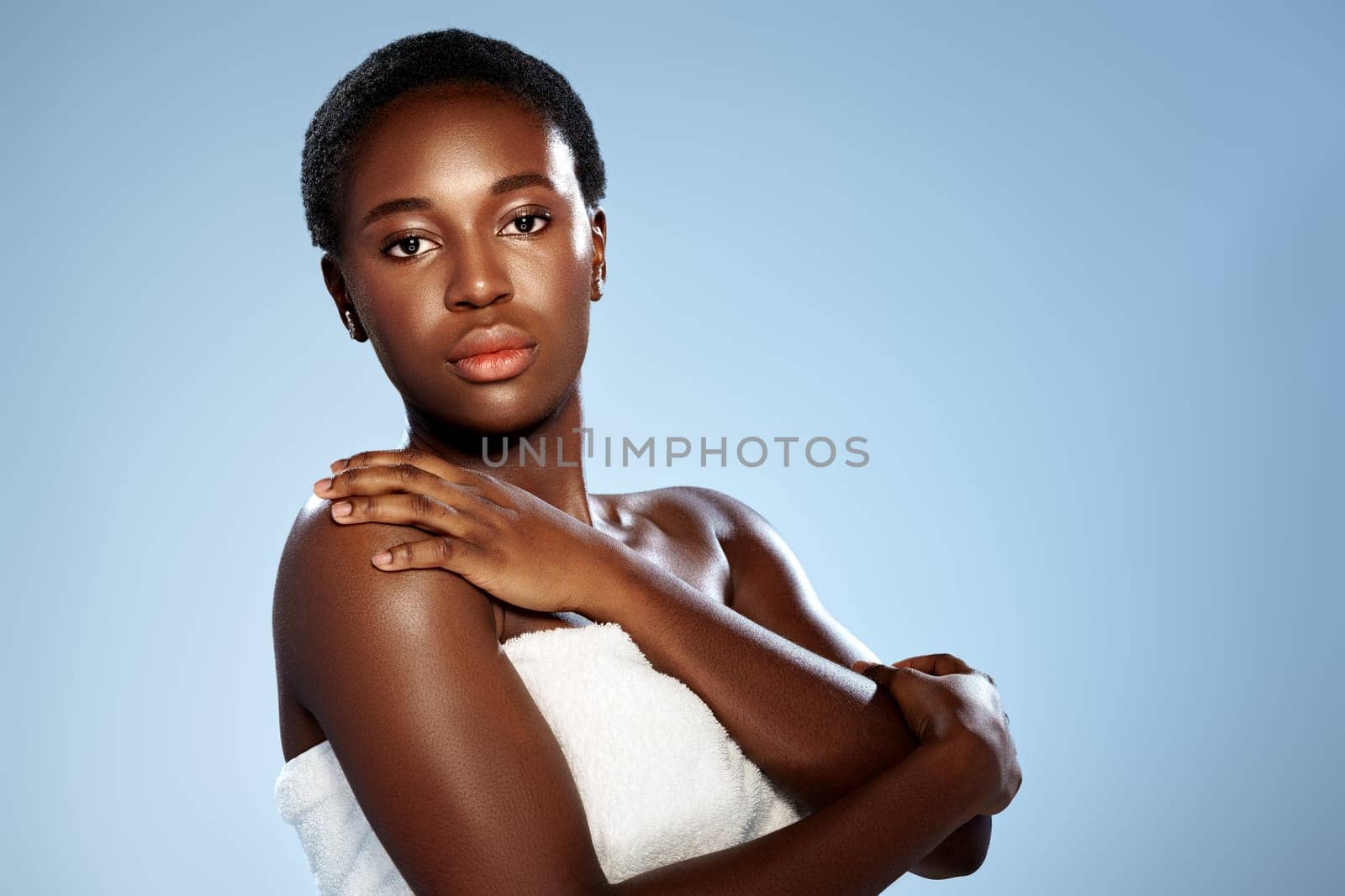 A woman with short dark hair poses confidently against a pale blue backdrop, her arms crossed, exuding grace and self-assurance. The soft lighting highlights her radiant skin.