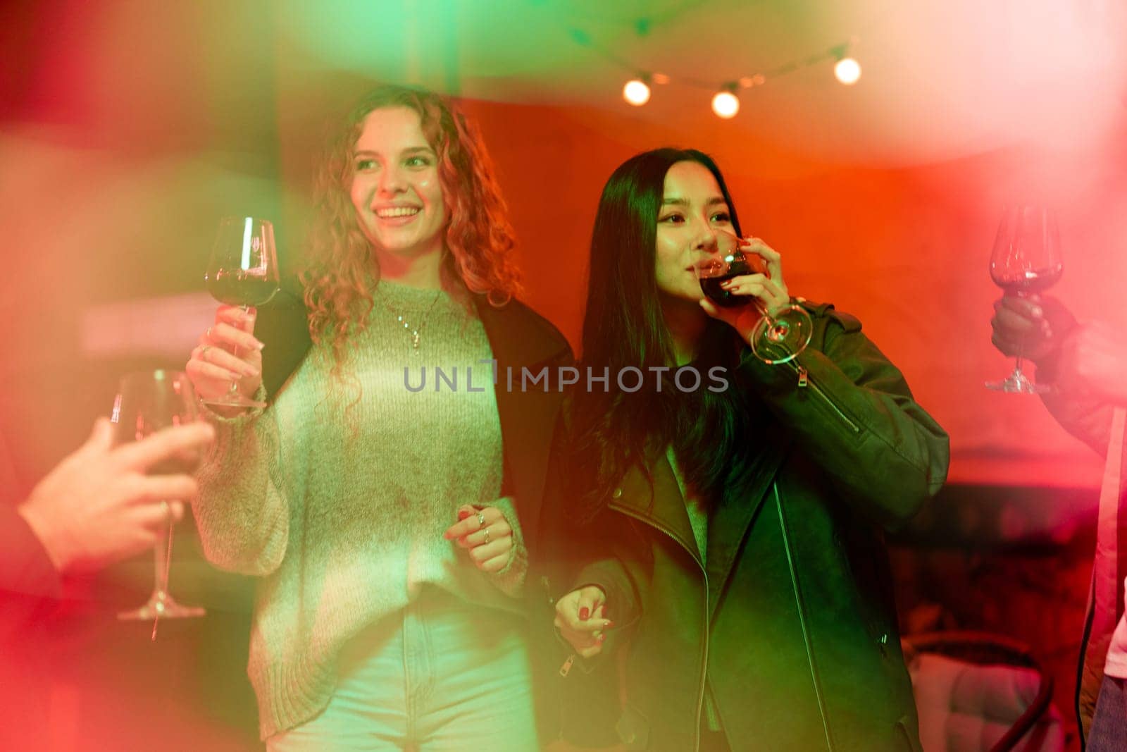 Two friends raise their glasses while laughing and enjoying a fun evening together at a cozy indoor gathering filled with colorful lights and good energy.
