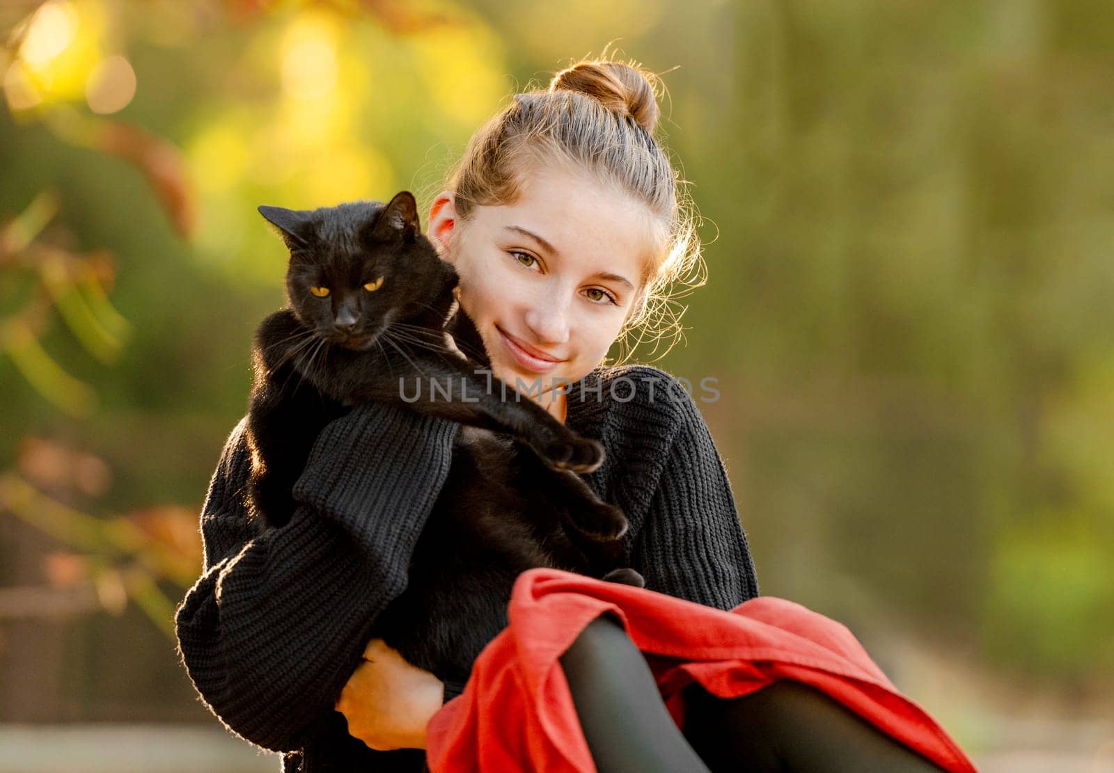 Pretty girl in red skirt hugging black cat outdoors at street with autumn leaves. Beautiful model teenager sitting with feline animal at park