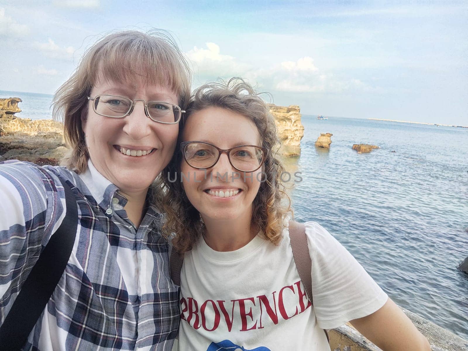Two friends capture a joyful selfie by the seaside, enjoying their travel adventure in a picturesque coastal location under a clear sky.