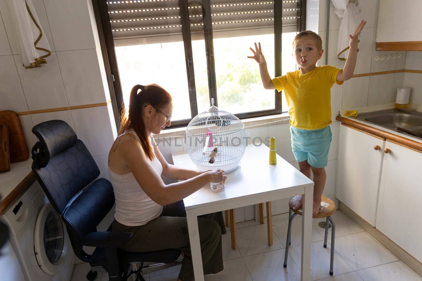 Mother taking care of pet bird in cage while her son is playing and making funny faces
