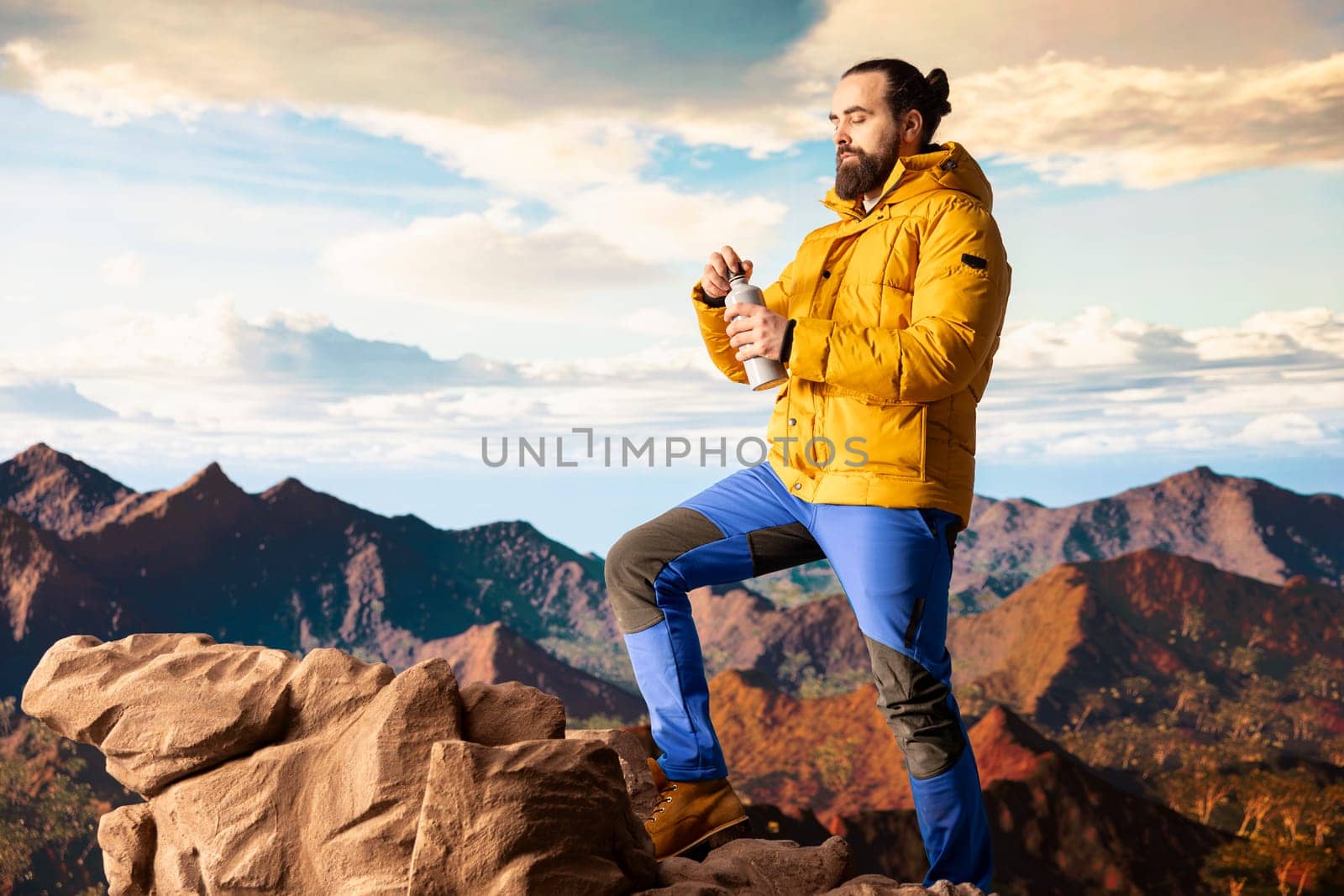 Young man explorer climbing the mountain range and hydrating with his water bottle, drinking refreshment on top of the rocky mountain. Male hiker refreshing after the trekking activity.