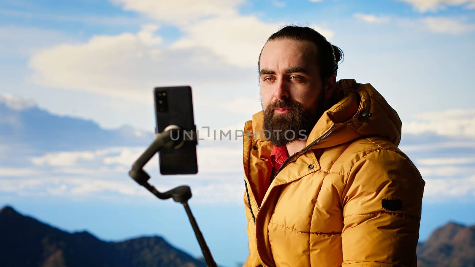 Traveler filming his hiking experience in the wilderness documenting his adventure on social media using his smartphone. Recording the beautiful scenic mountain view behind him for content. Camera B.