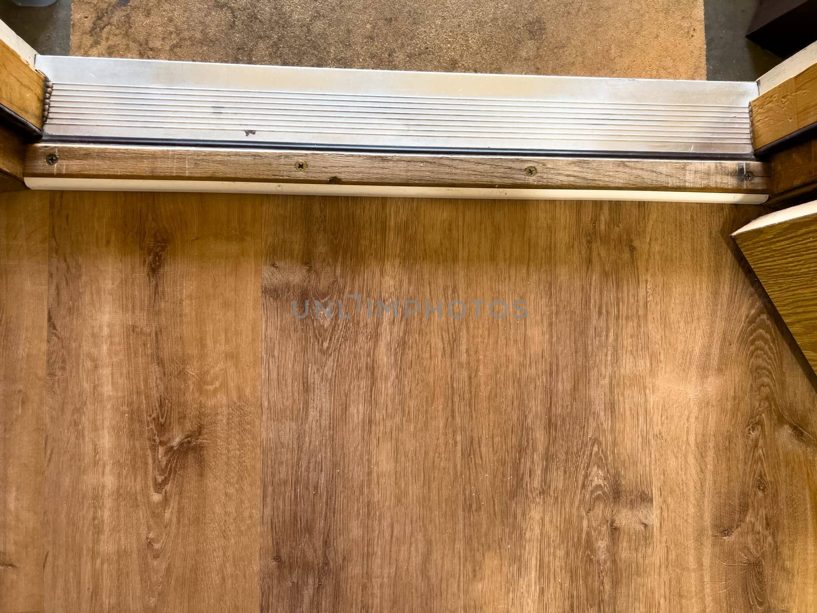 Close-up view of an interior door threshold showing the transition between wood flooring and a concrete surface. The metal strip and surrounding frame highlight the textures and construction details of a residential entryway.