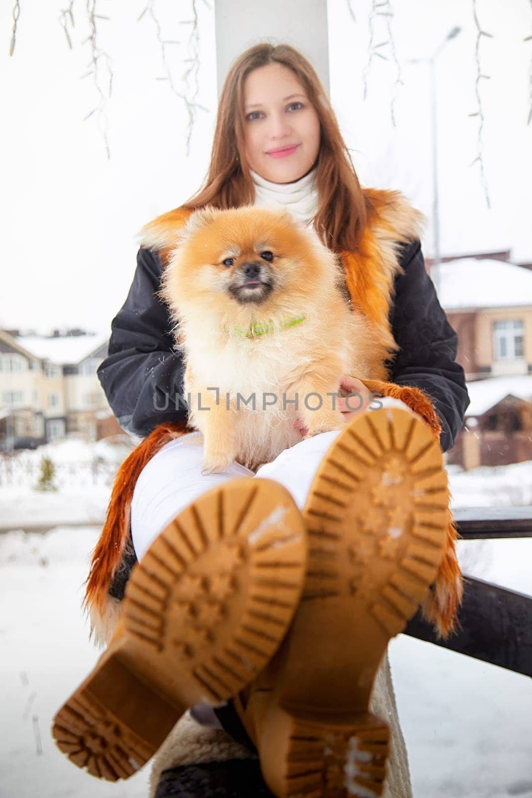 A woman lovingly holds her fluffy Pomeranian while seated on a snowy bench in a residential area.