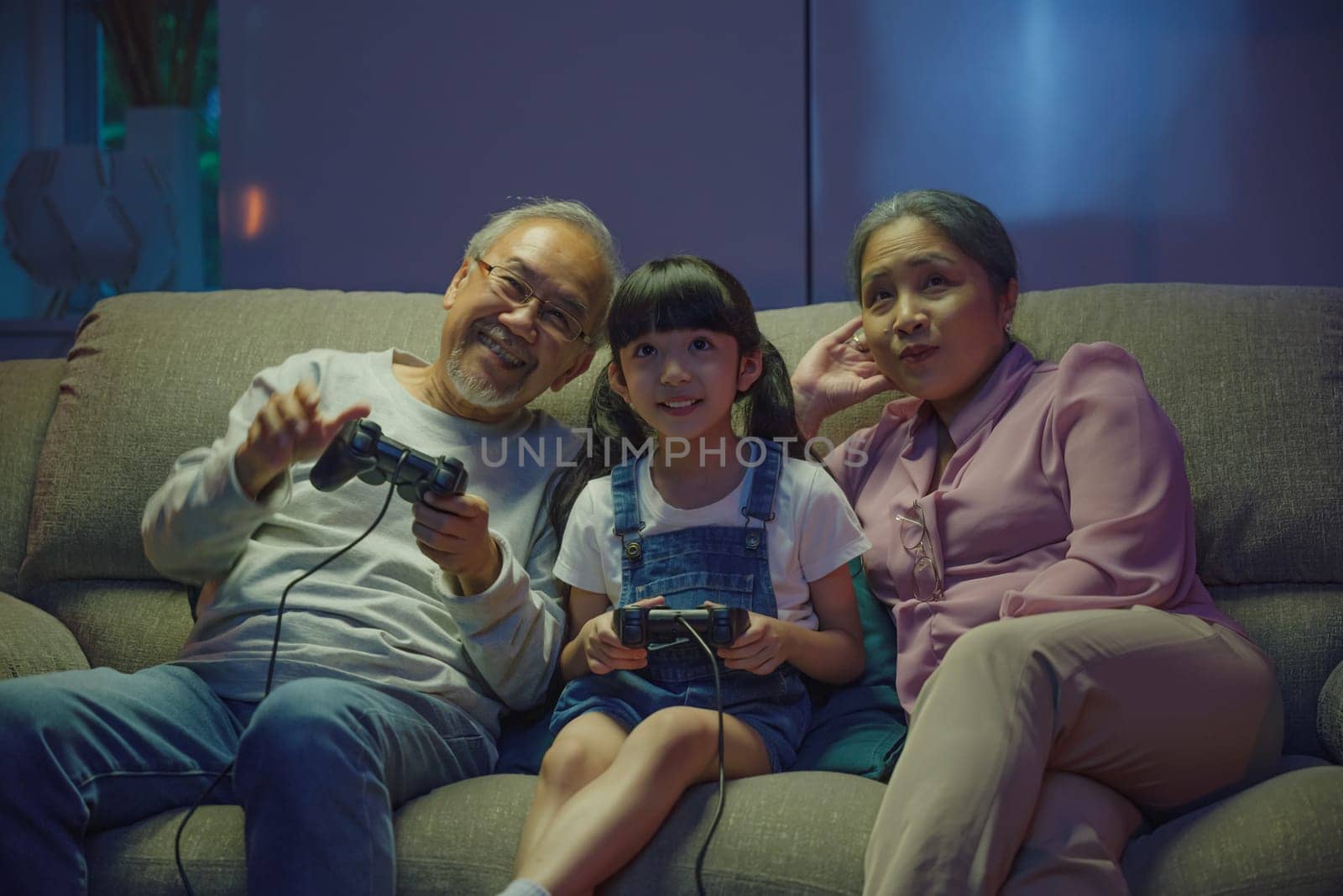 Happy Asian family bonding over a video game on a cozy couch, little girl playing video games console with senior grandparents together with controller joysticks while sitting on the sofa at night