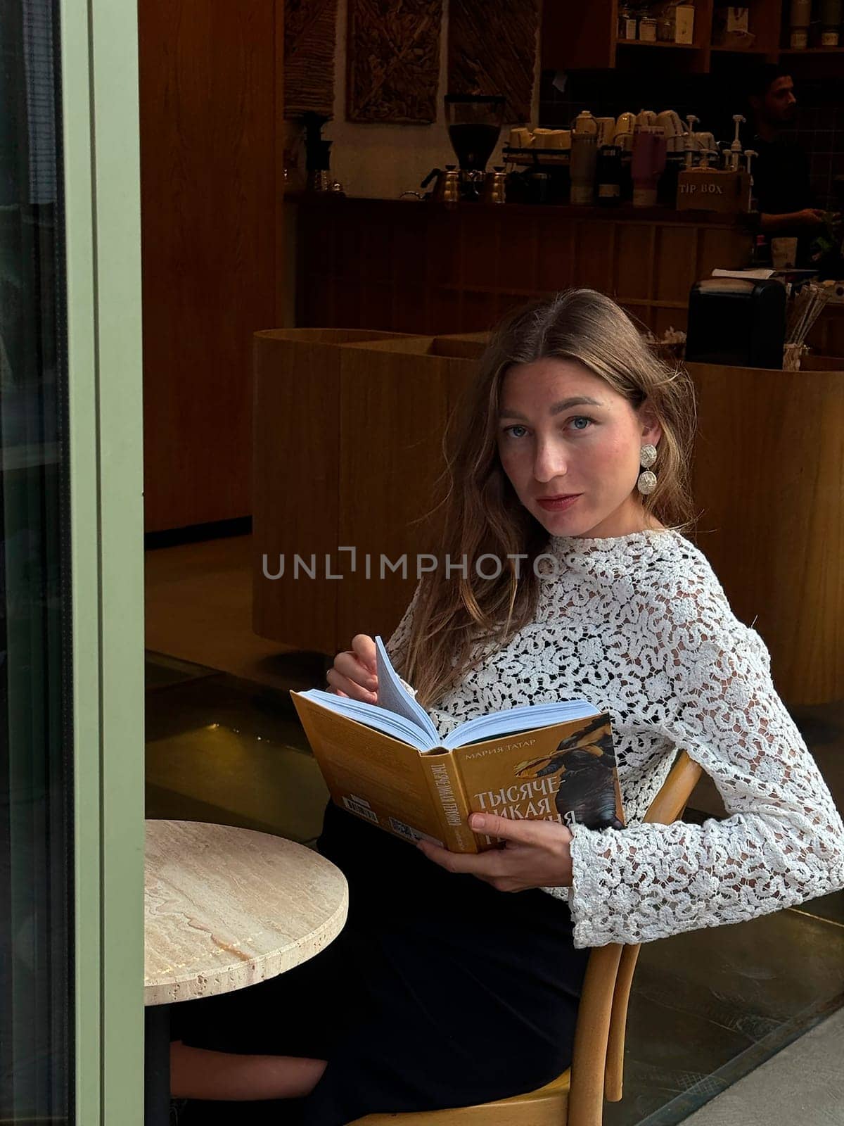 A young woman sits at a cafe table, reading a book while dressed in a stylish outfit. The atmosphere is warm and inviting.