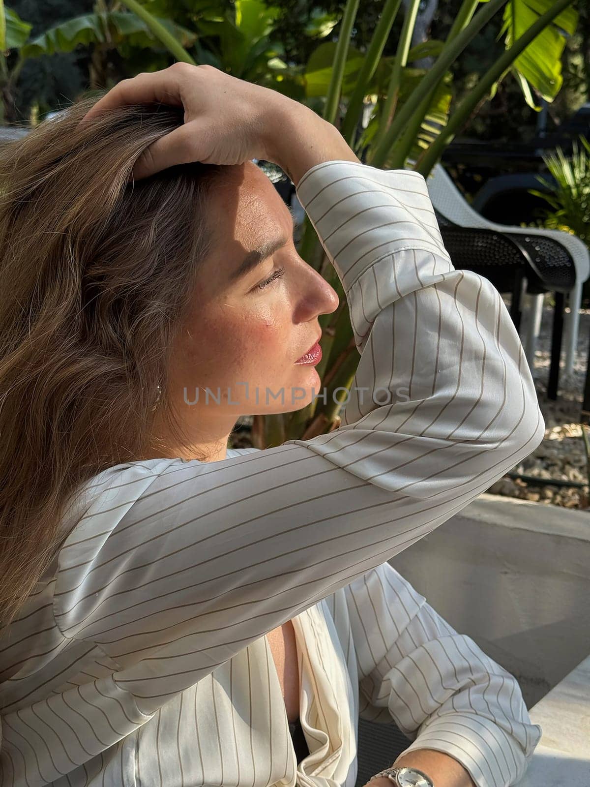 A woman sits thoughtfully in a cafe surrounded by greenery, enjoying the warm afternoon light and taking a break.