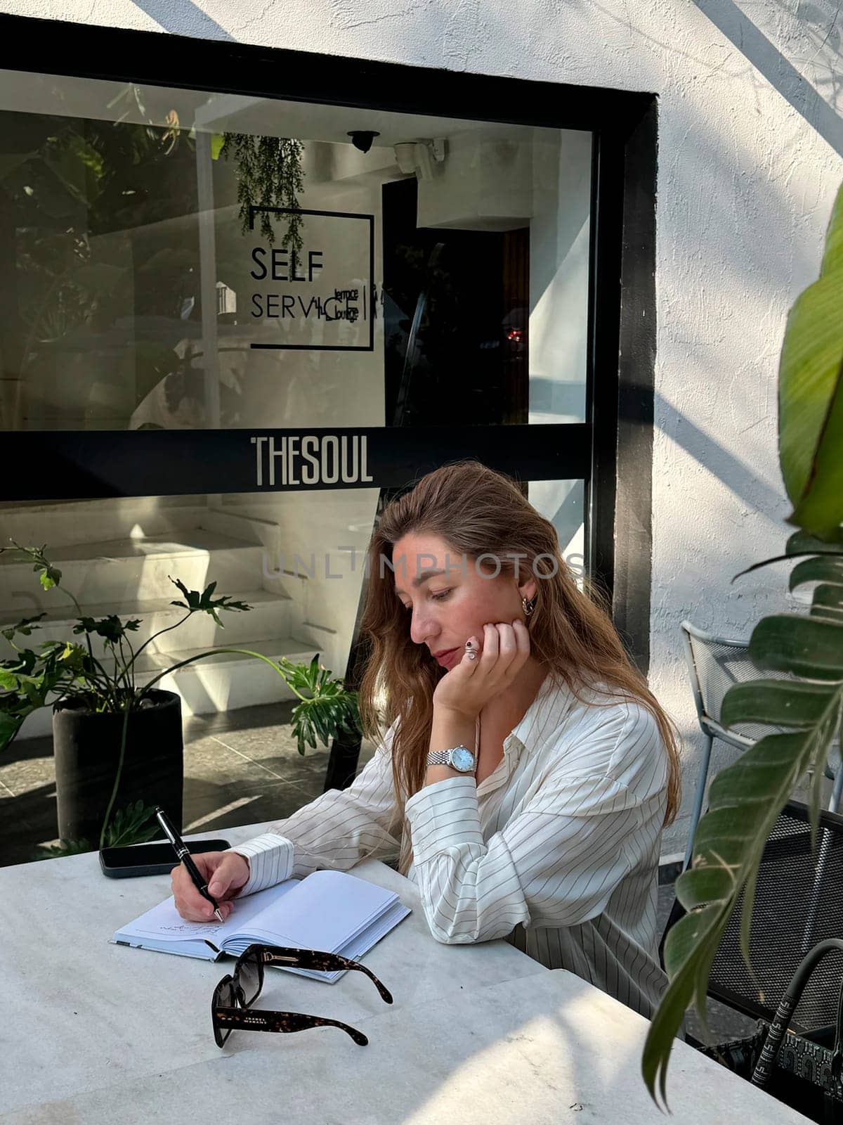 A woman sits at a table in a cafe surrounded by greenery, focused on writing in her notebook, enjoying the peaceful atmosphere.