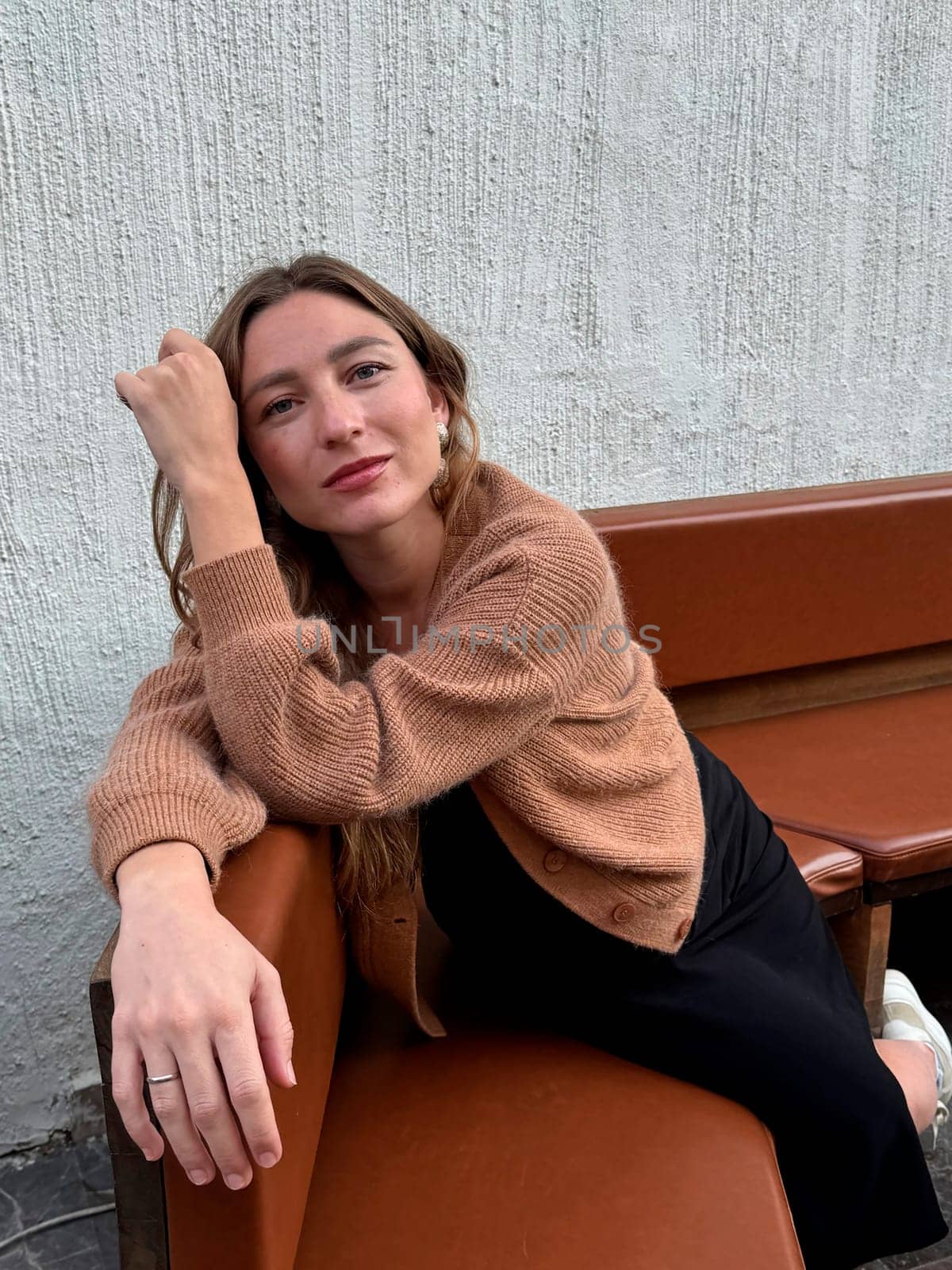 A young woman leans comfortably on a brown bench, showcasing a relaxed pose, in a bright urban environment.