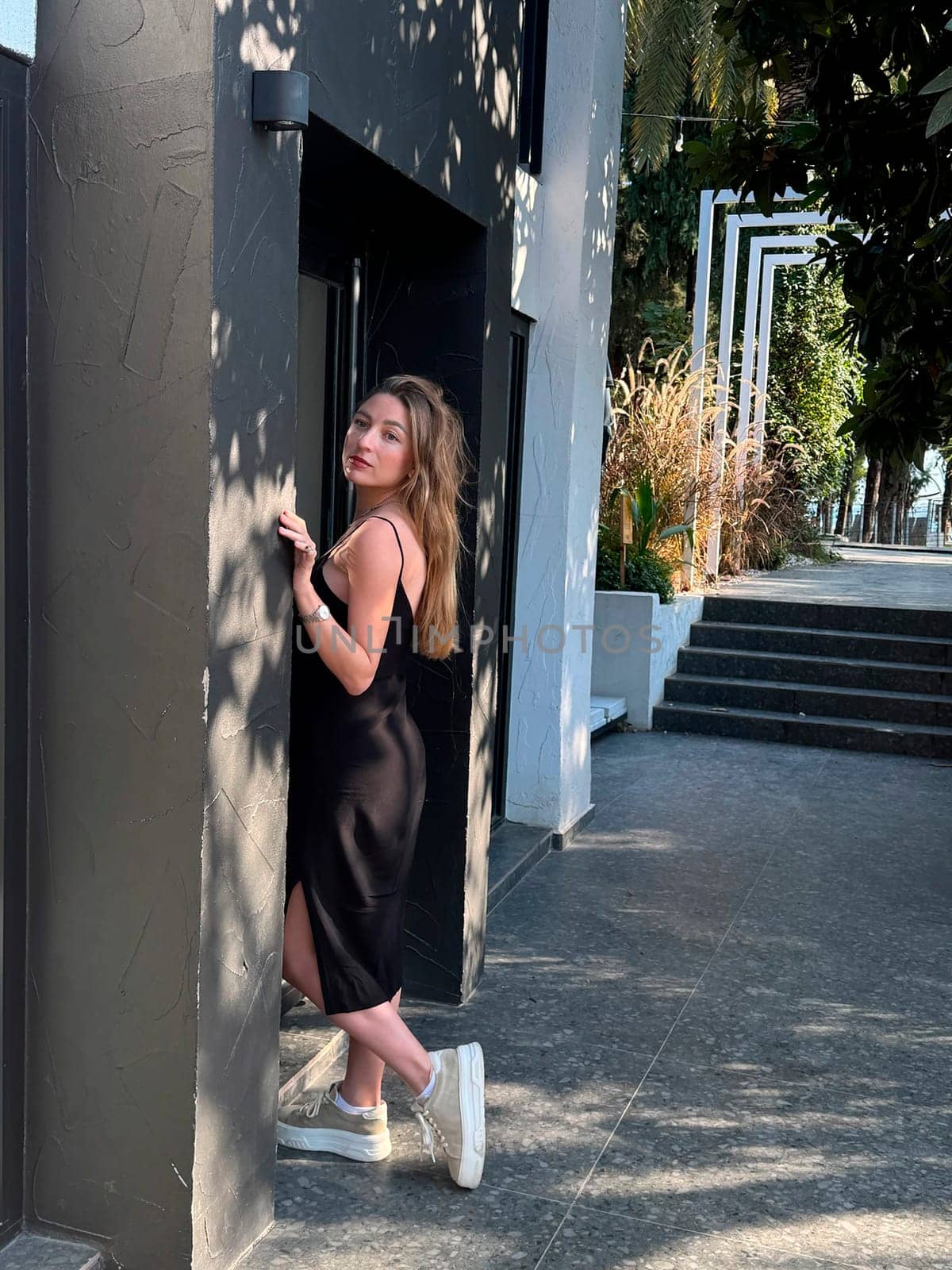 A woman stands against a stylish wall, showcasing her relaxed style in a black dress and sneakers.