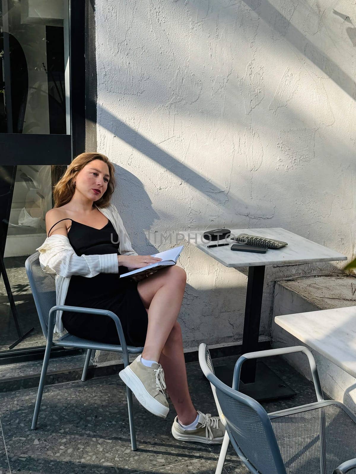 A woman sits comfortably at a cafe table, writing in a notebook, enjoying the warm afternoon sun. She wears a casual outfit and appears focused.