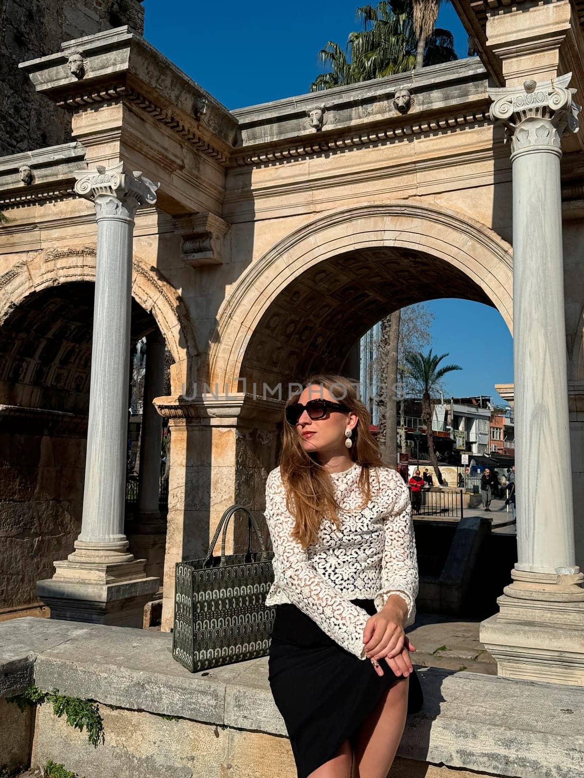 A stylish woman sits confidently on a stone structure, enjoying the sun and showcasing her chic outfit near historic ruins.