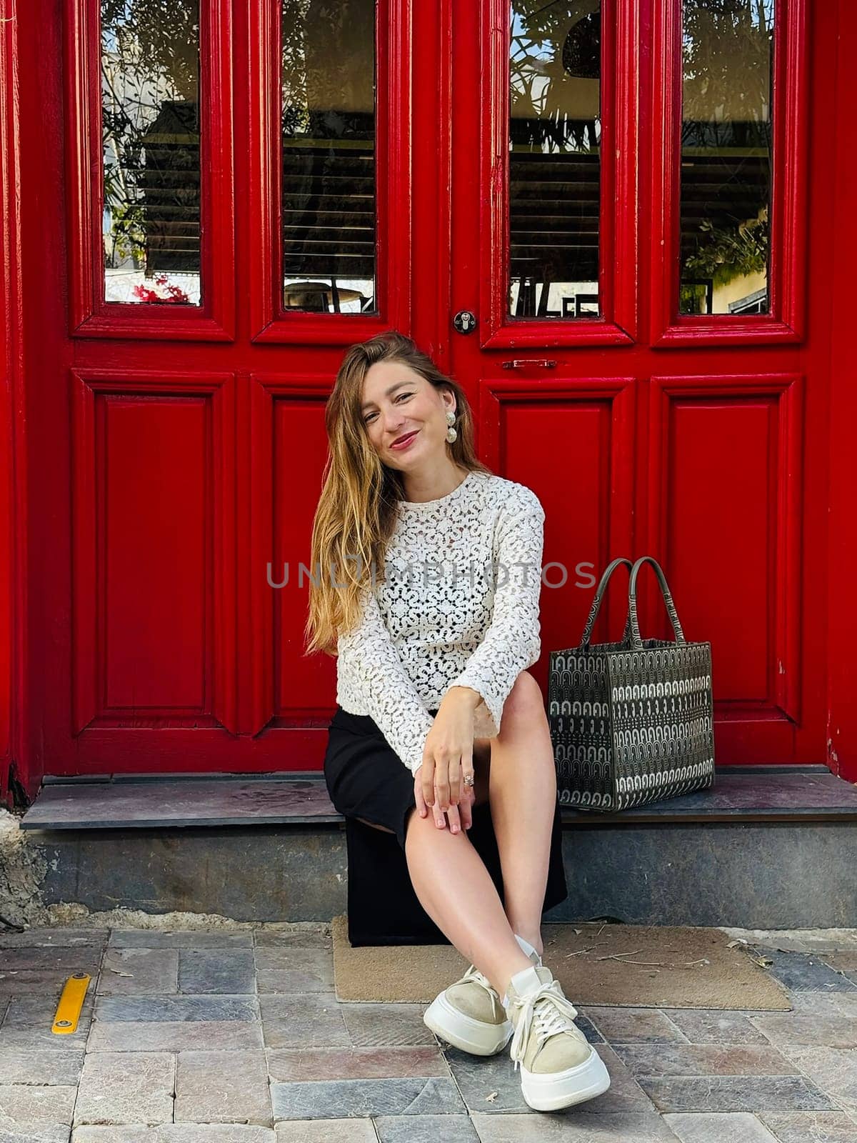 A young woman relaxes on steps near a bright red door, dressed in a stylish outfit, enjoying the warm day.