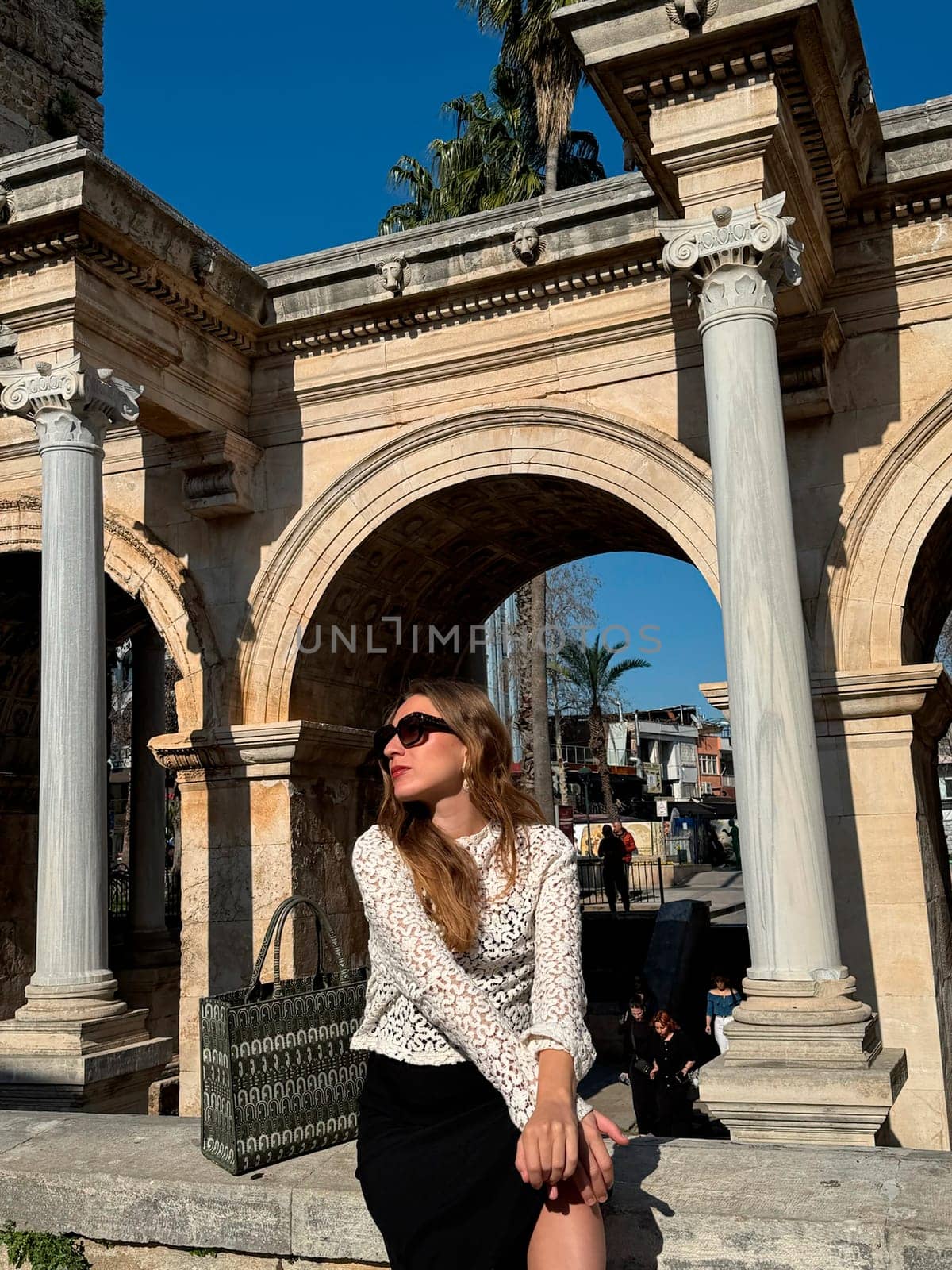 A young woman relaxes under grand arches in a historic location, surrounded by palm trees and lively atmosphere during daytime.