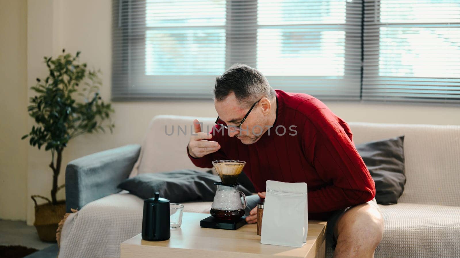 Older man in red sweater smelling coffee brewed with pour-over set on wooden table.