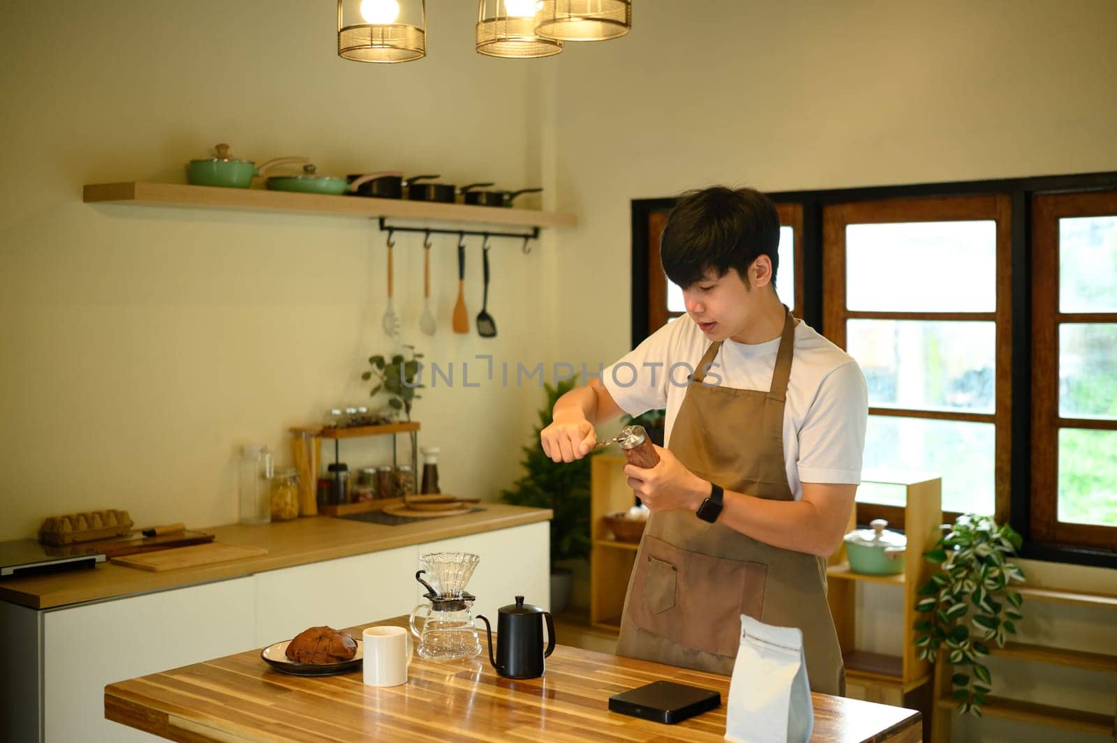 Young man in apron grinding fresh coffee beans in a home kitchen.
