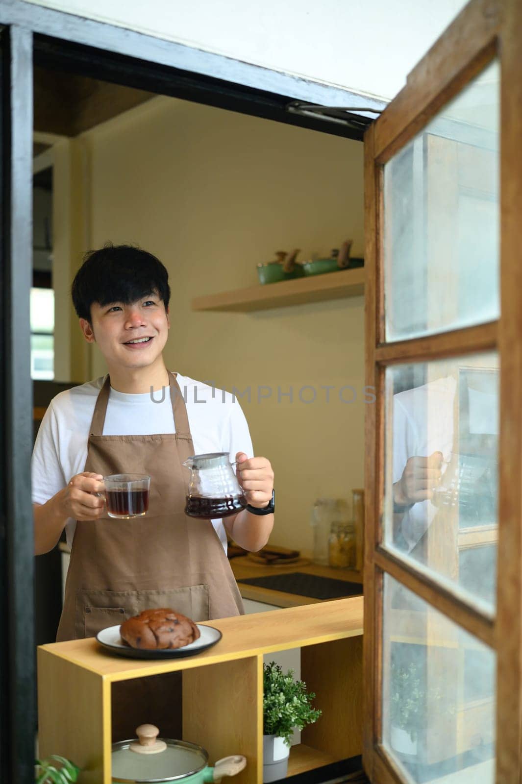 Young man holding a fresh cup of coffee and carafe, smiling through a window.