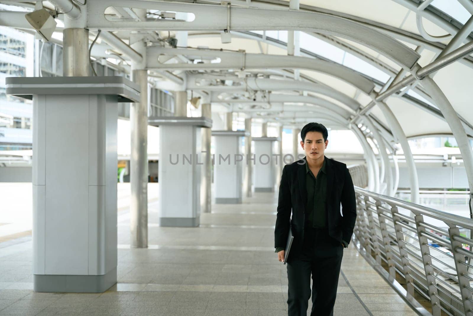 Confident businessman walking through a modern city walkway with a laptop in hand.