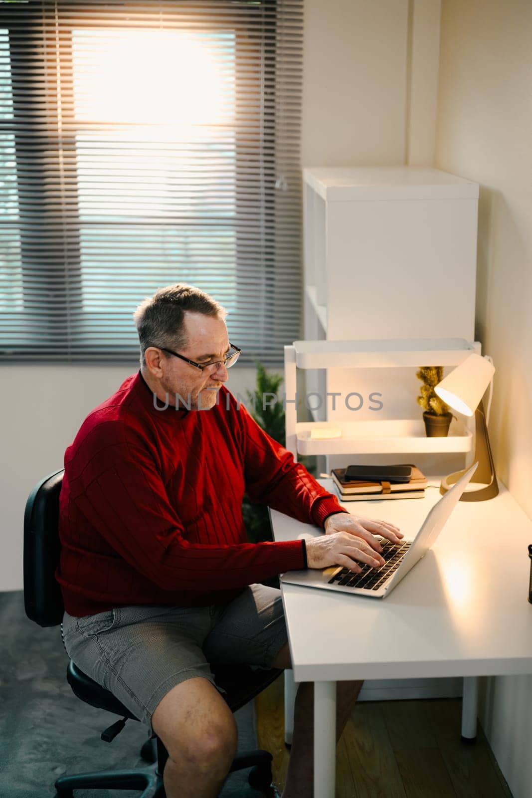 Senior man in red sweater working on a laptop at minimalist desk under desk lamp.