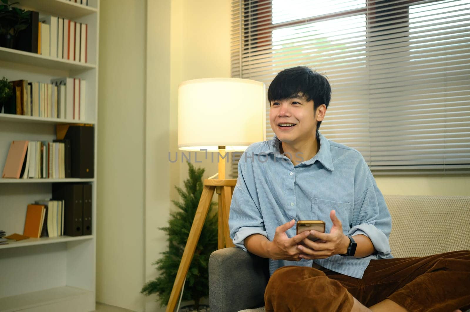 Charming young adult male sitting in cozy living room, using mobile phone.