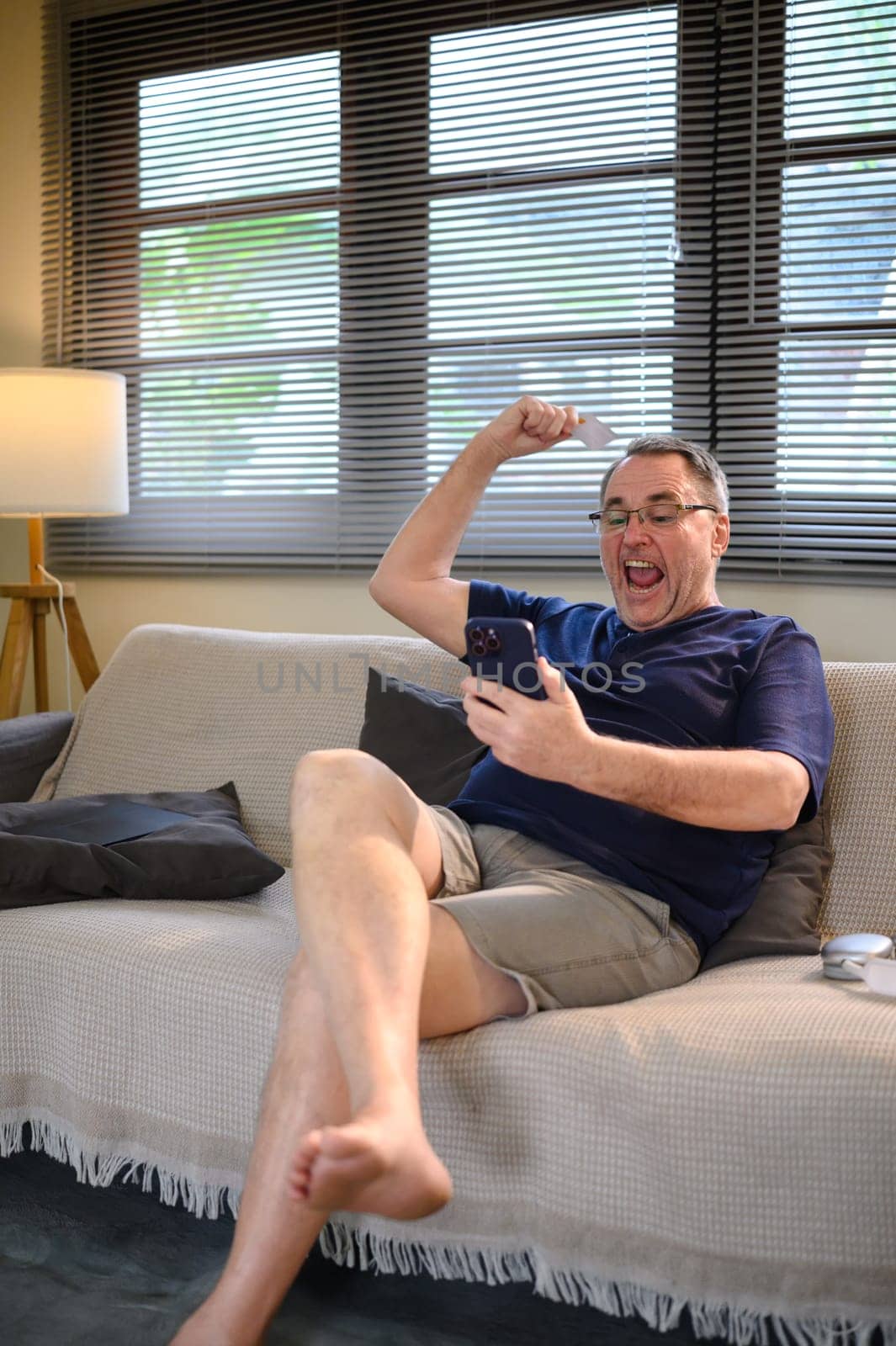 Excited senior man celebrating online success while holding smartphone and credit card.