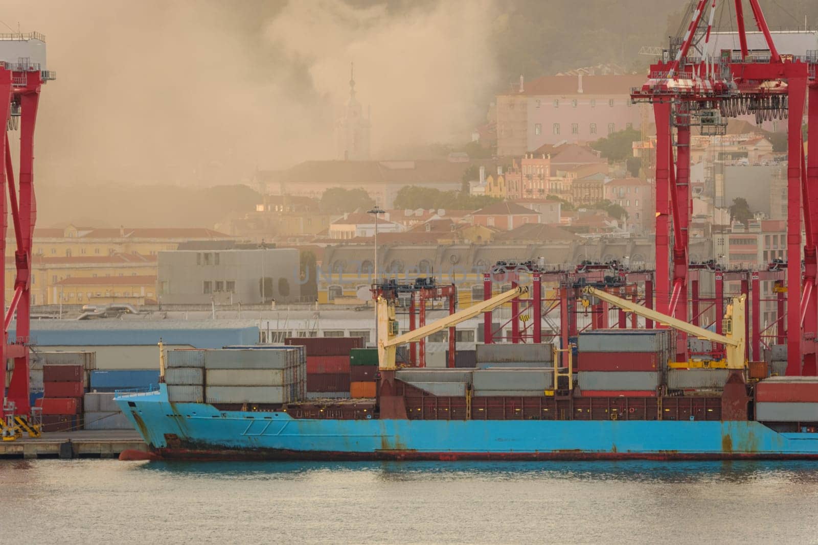 Blue container ship docked at Lisbon port, red cranes and stacks of shipping containers on deck, city buildings and church spire in evening haze and fire smoke in background. Risk of fires in summer, drought and heatwave, danger, damage, Portugal