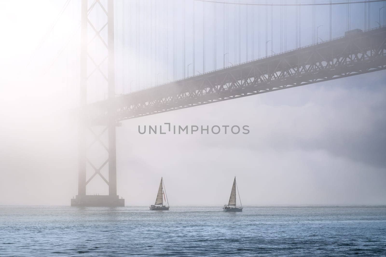 Dramatic view of Lisbon iconic 25 de Abril bridge in morning fog on sunny day, two sailboats navigate calm Tagus river, Portugal. Misty atmosphere, peaceful, quiet scene, mist roll in over waters, riverscape, suspension bridge, beauty in nature.