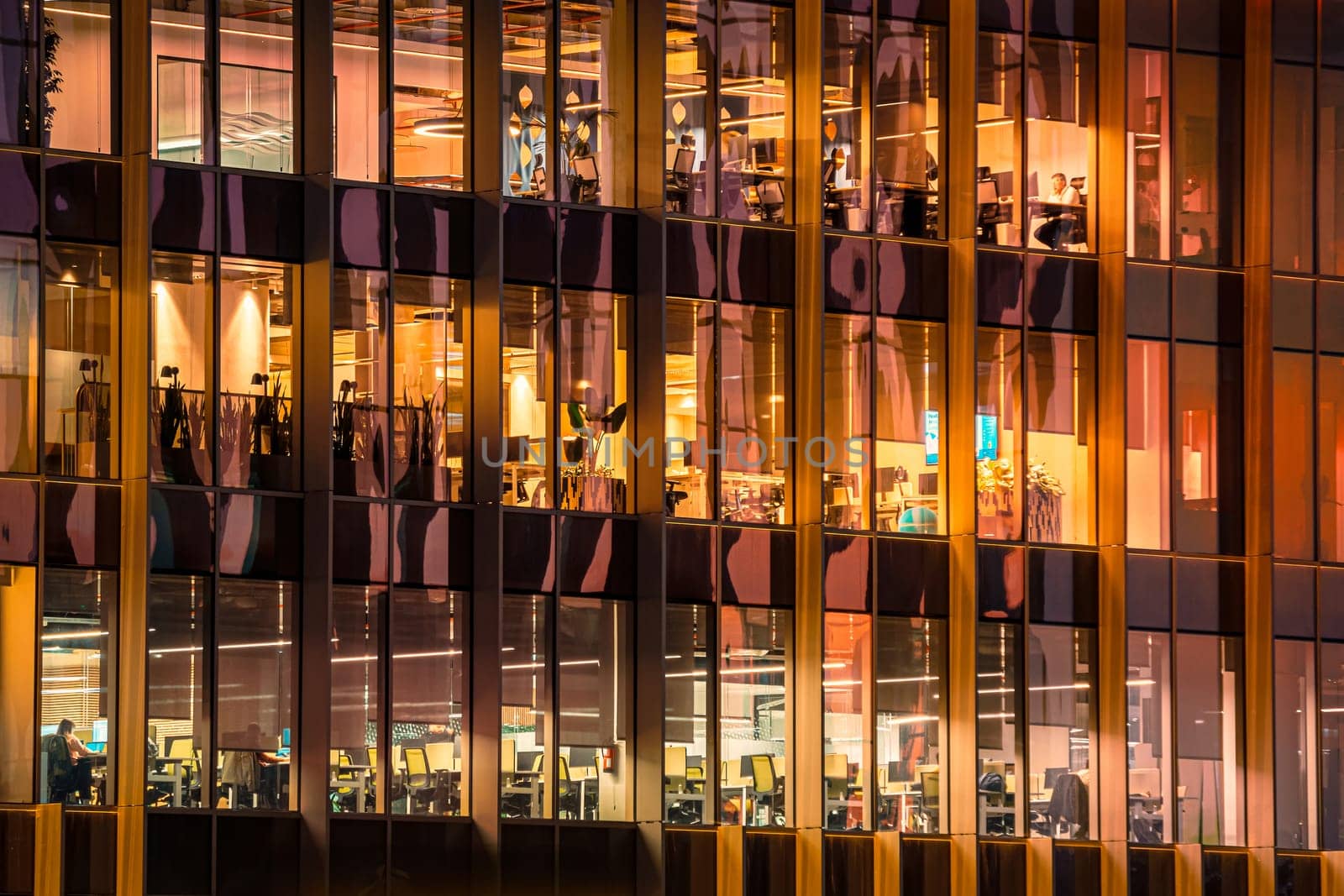 Contemporary office building at night with large glass windows revealing warmly lit workspaces. Employees are visible at desks and computers, active business atmosphere and modern corporate environment, after sunset, modern work environment
