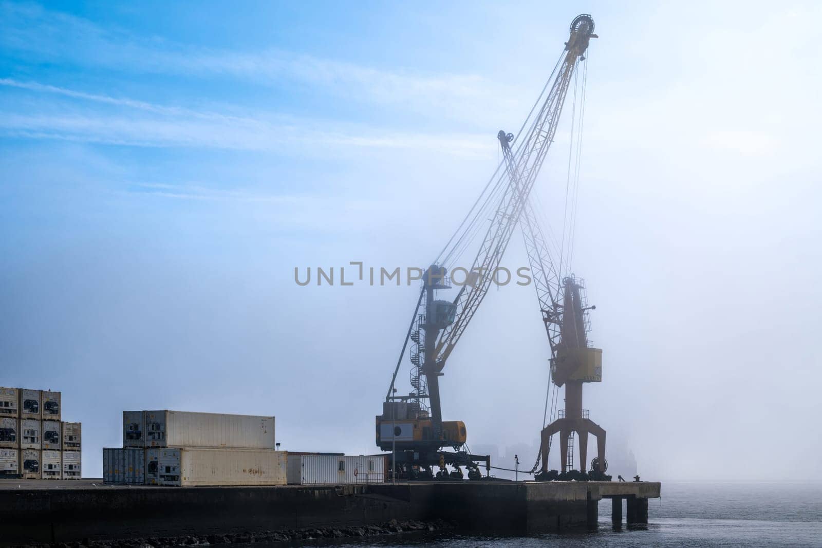 Big industrial cranes on pier beside stacked shipping containers in morning fog. Misty waterfront scene highlight maritime industry, logistics and atmosphere of busy cargo port at dawn on sunny day. Cargo operations, docks, clear blue sky, silhouette
