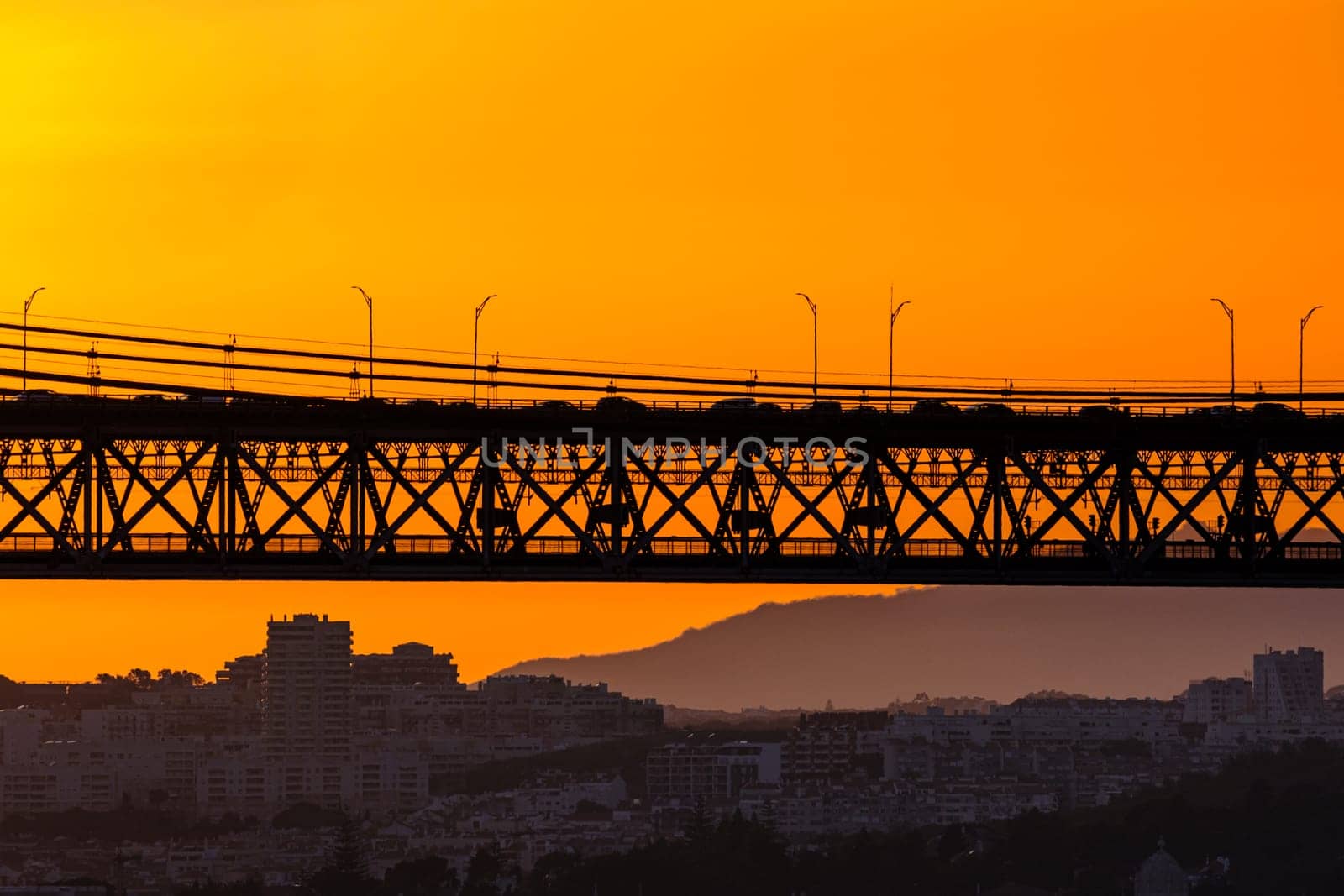 Bridge spans city skyline, silhouetted against vivid orange sunset. Soft city outlines and distant hills frame flowing late evening traffic, dynamic urban energy, tranquil evening light for unique harmony of architecture and nature. Urban landscape