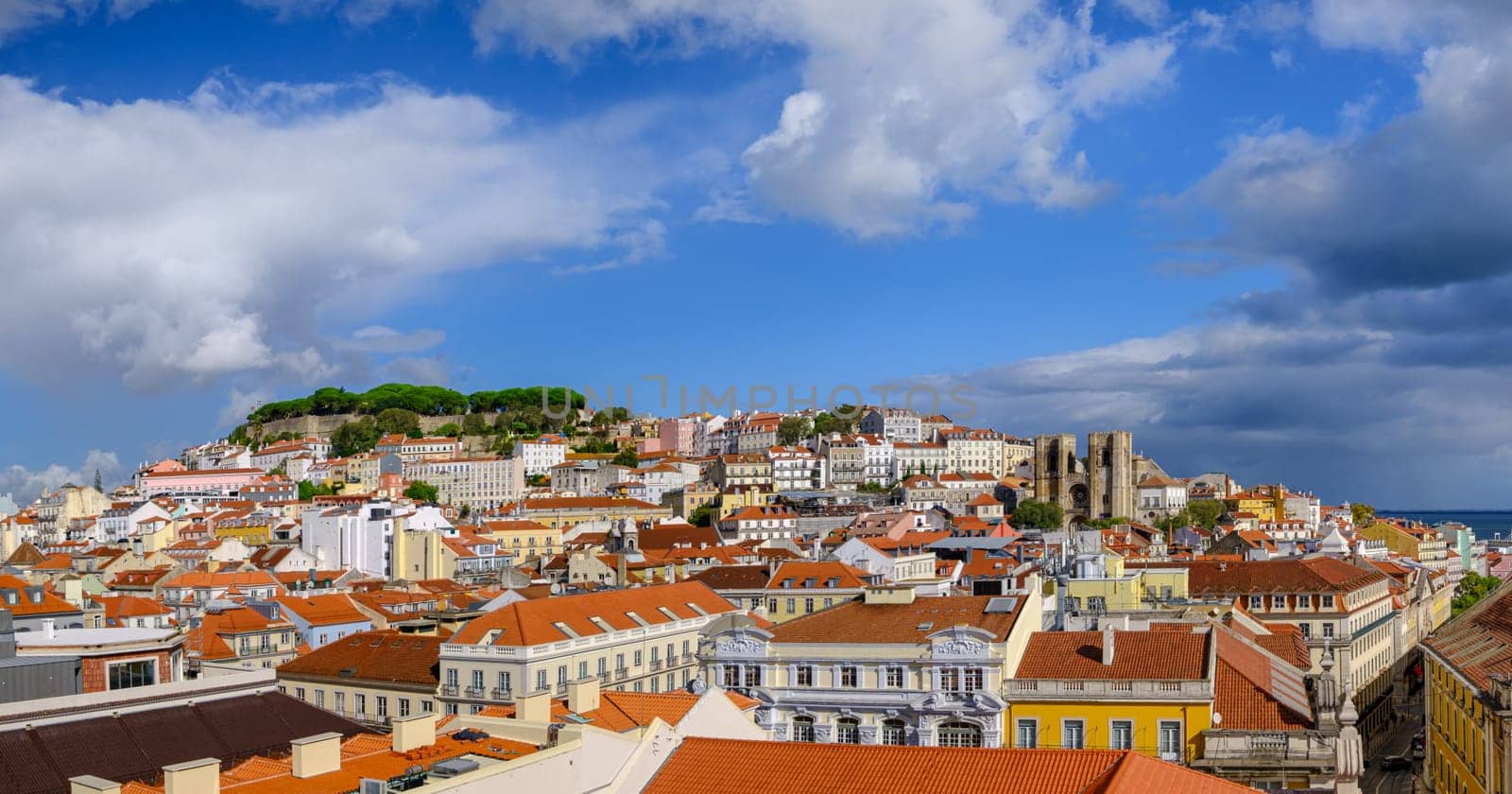 Scenic panoramic view of Lisbon historic Alfama district, St George castle on hill top with lush foliage, Gothic cathedral Se and colorful houses with terracotta rooftops on sunny day, clear blue sky and dramatic clouds. Famous travel destination.