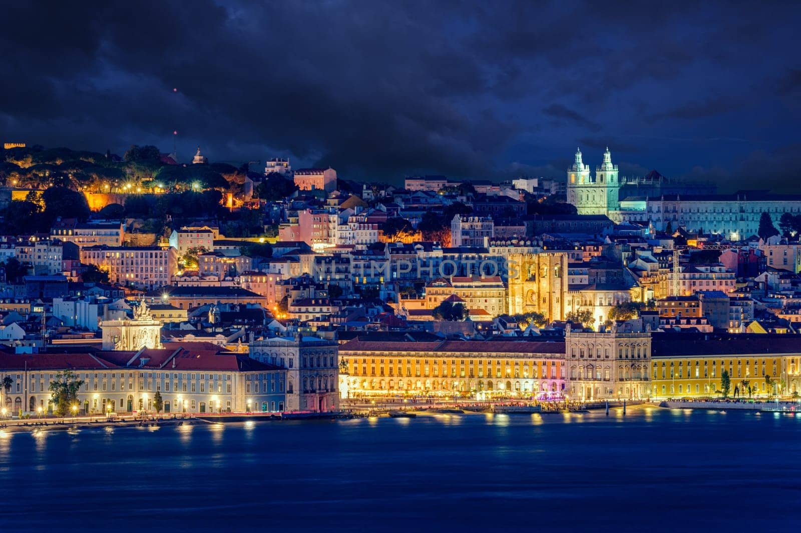 Lisbon historic old town with vibrant city lights under dramatic night sky. Praca do Comercio or Commerce square and iconic landmarks by waterfront of Tagus river, cityscape after dark, churches and cathedrals, scenic view of hilltop castle Sao Jorge