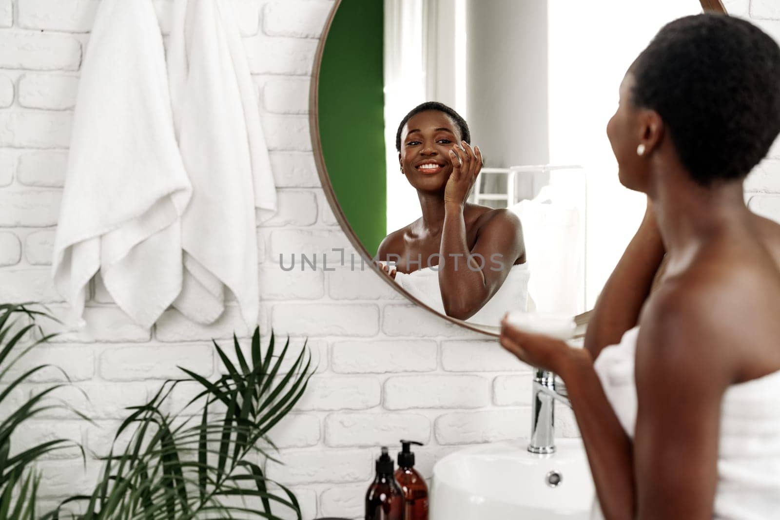A woman wrapped in a towel smiles while applying skincare in a stylish bathroom. The setting features green accents, a mirror, and potted plants, creating a serene atmosphere.