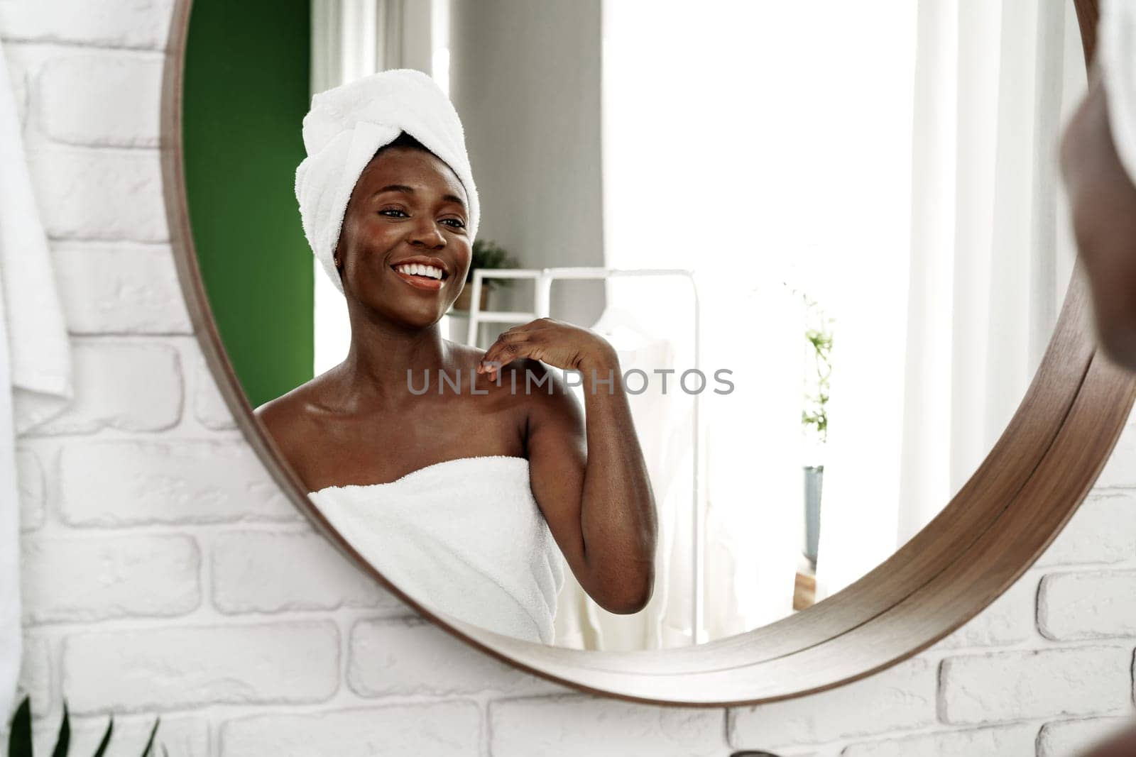 A woman enjoys a moment of self-care in the bathroom, smiling confidently at her reflection in a round mirror. The bright setting adds to her joyful expression.