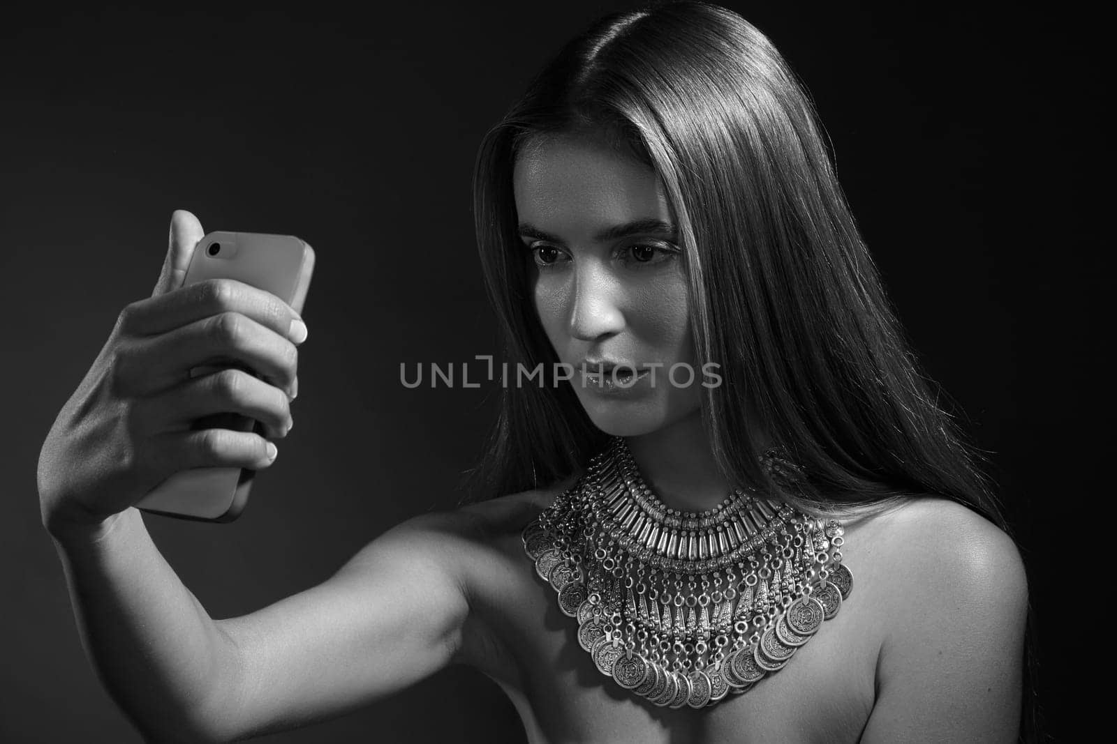 Beauty style portrait. Stylish young woman with straight hair taking a selfie with her smartphone, wearing a large Indian-style silver necklace. Concept of self-expression, influencer lifestyle, and ethnic fashion.