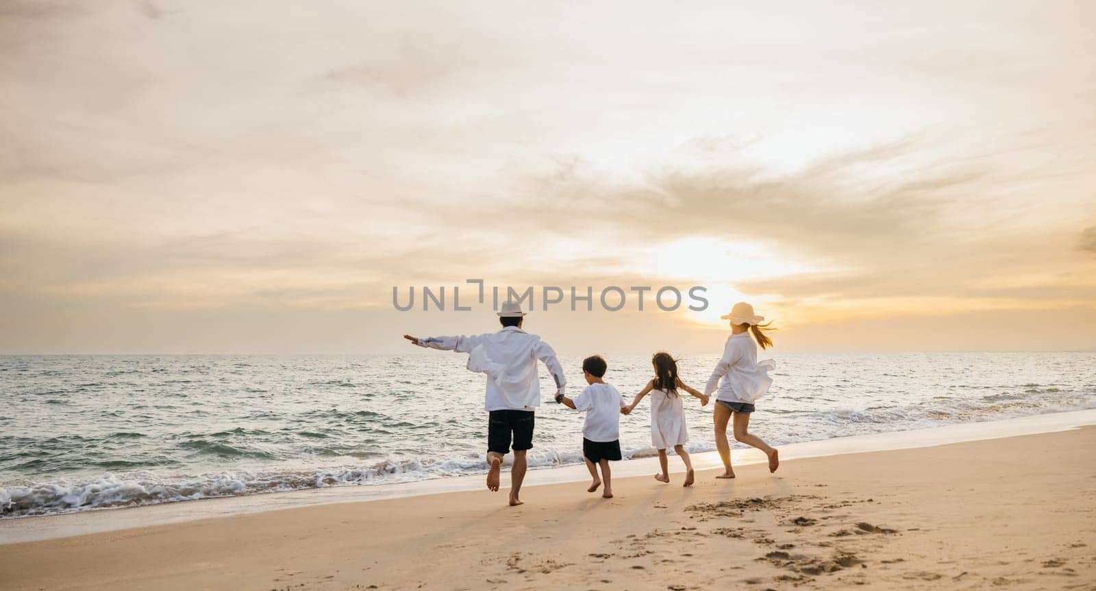 A happy Asian family seen from behind enjoys sunny day at beach. Parents and daughters bond running jumping and pretending to fly with arms outstretched. carefree family moment by sea.