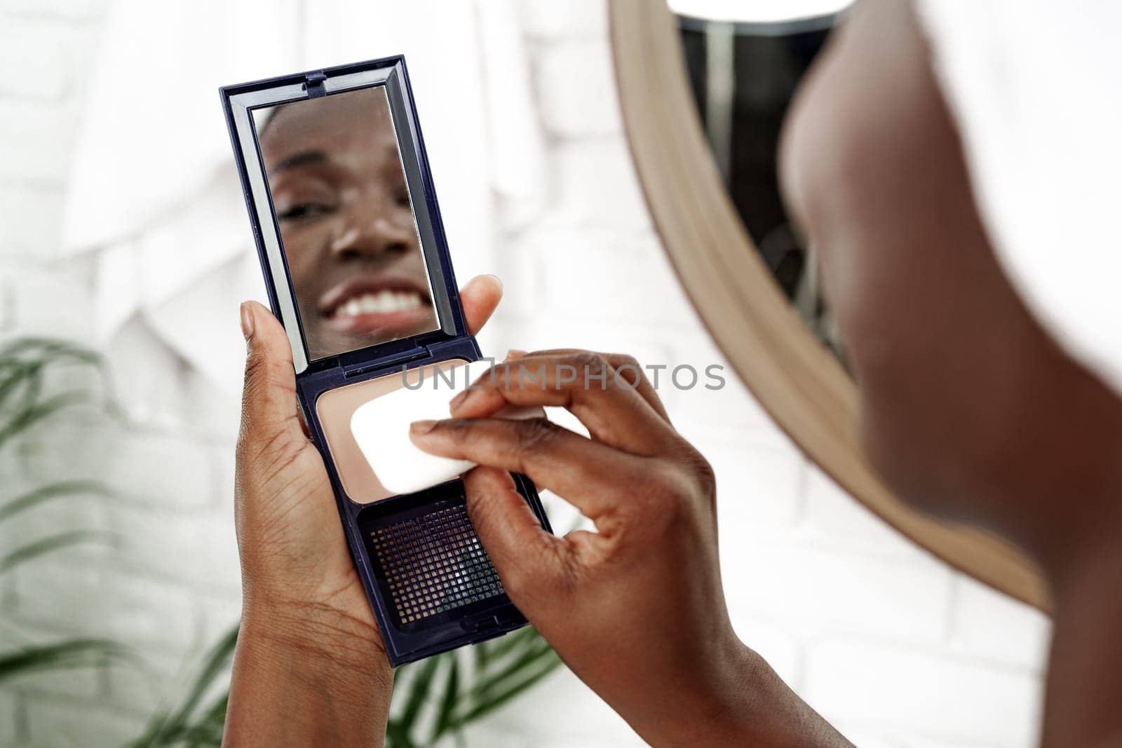 A woman is using a compact mirror in her bathroom to apply makeup. She is smiling while focusing on achieving a polished appearance as part of her daily routine.