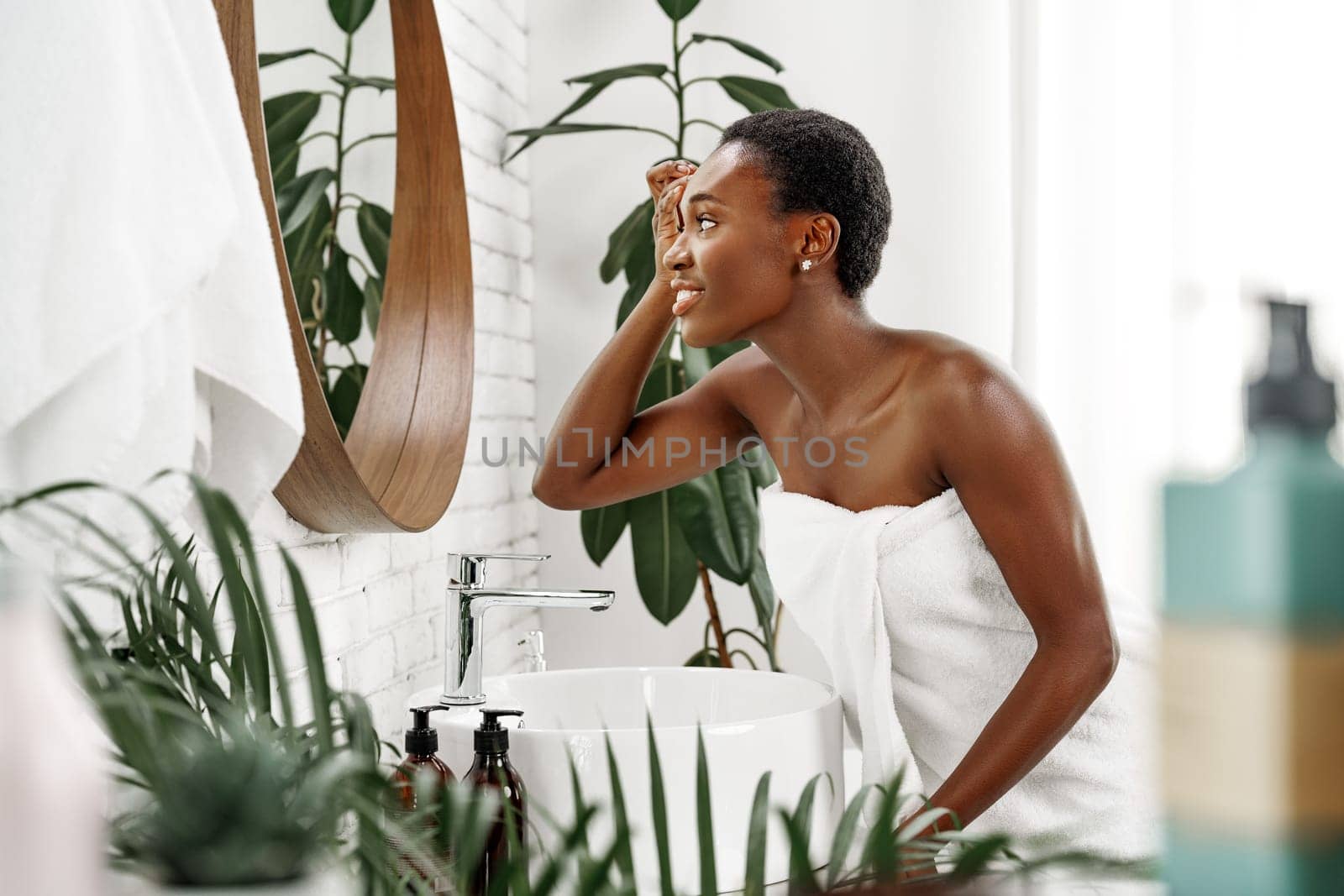A woman applies skincare products in a stylish bathroom filled with greenery. The sunlight highlights her careful routine while she enjoys a moment of self-care.