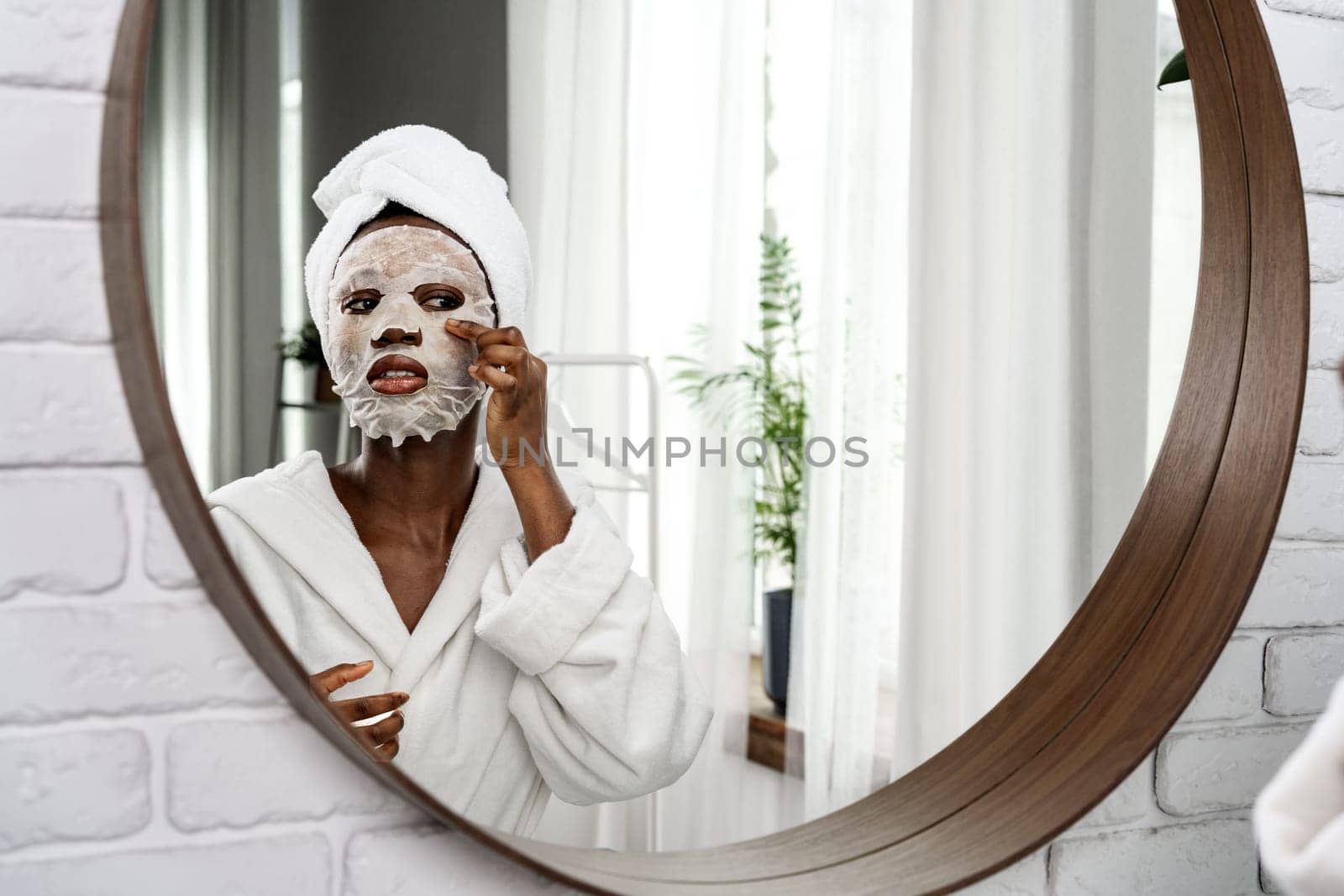 In a well-lit bathroom, a woman in a bathrobe applies a facial mask while looking at her reflection in a round mirror. Natural light creates a calming atmosphere.