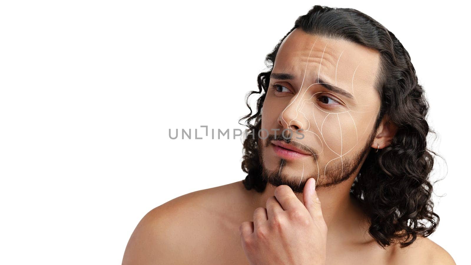 A young man with curly hair and a beard contemplates his skin's appearance, showcasing subtle facial lines and texture under bright lighting in a plain background.