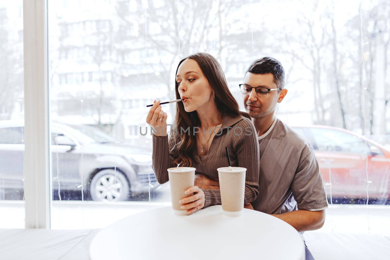 A couple is seated at a round table in a cafe while the snow falls outside. The man gently holds the woman from behind as she tastes a sweet treat with a stick.