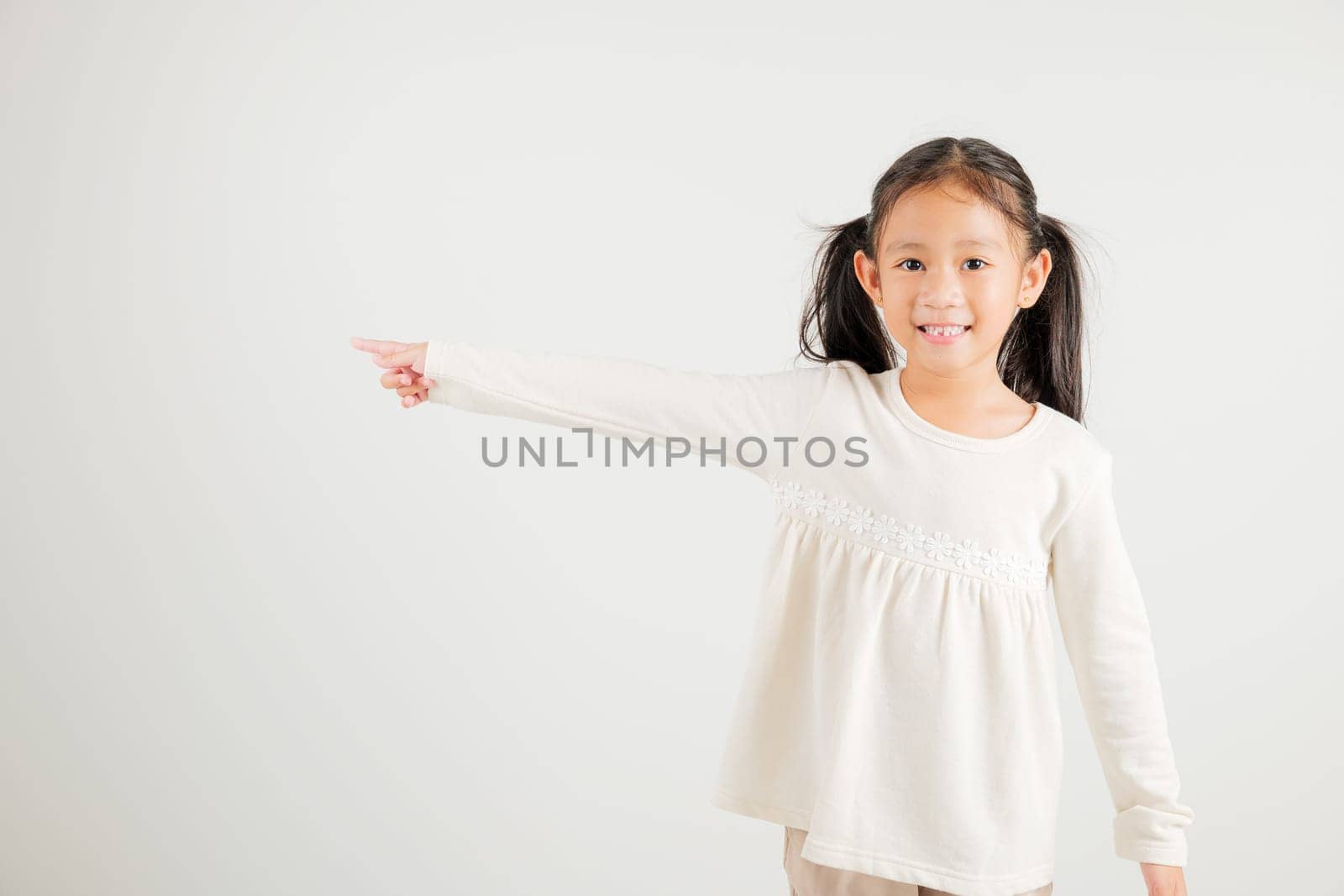 Happy Asian portrait cute young kid girl smiling indicate finger empty space, studio shot isolated on white background, Thai kindergarten child pointing index out with copy space