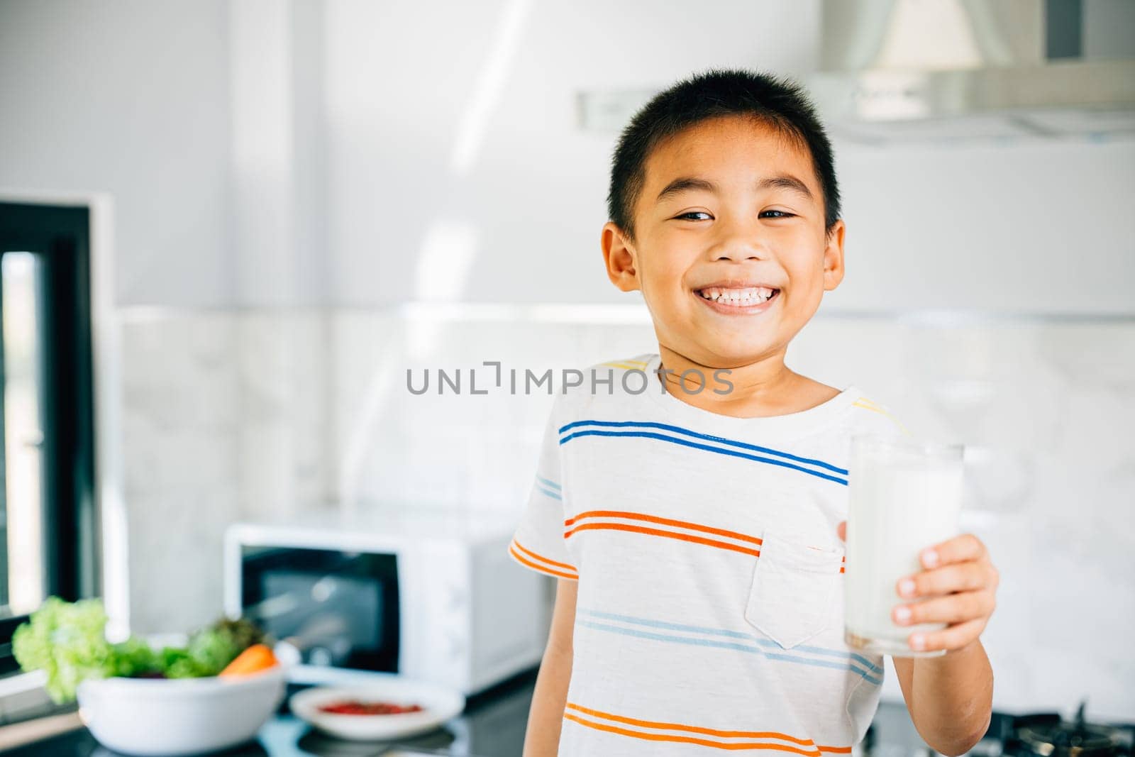 Portrait of adorable Asian preschooler, boy holds milk in kitchen. Smiling son enjoys drink, radiating joy. Happy child savoring calcium-rich liquid, feeling cheerful at home give me.