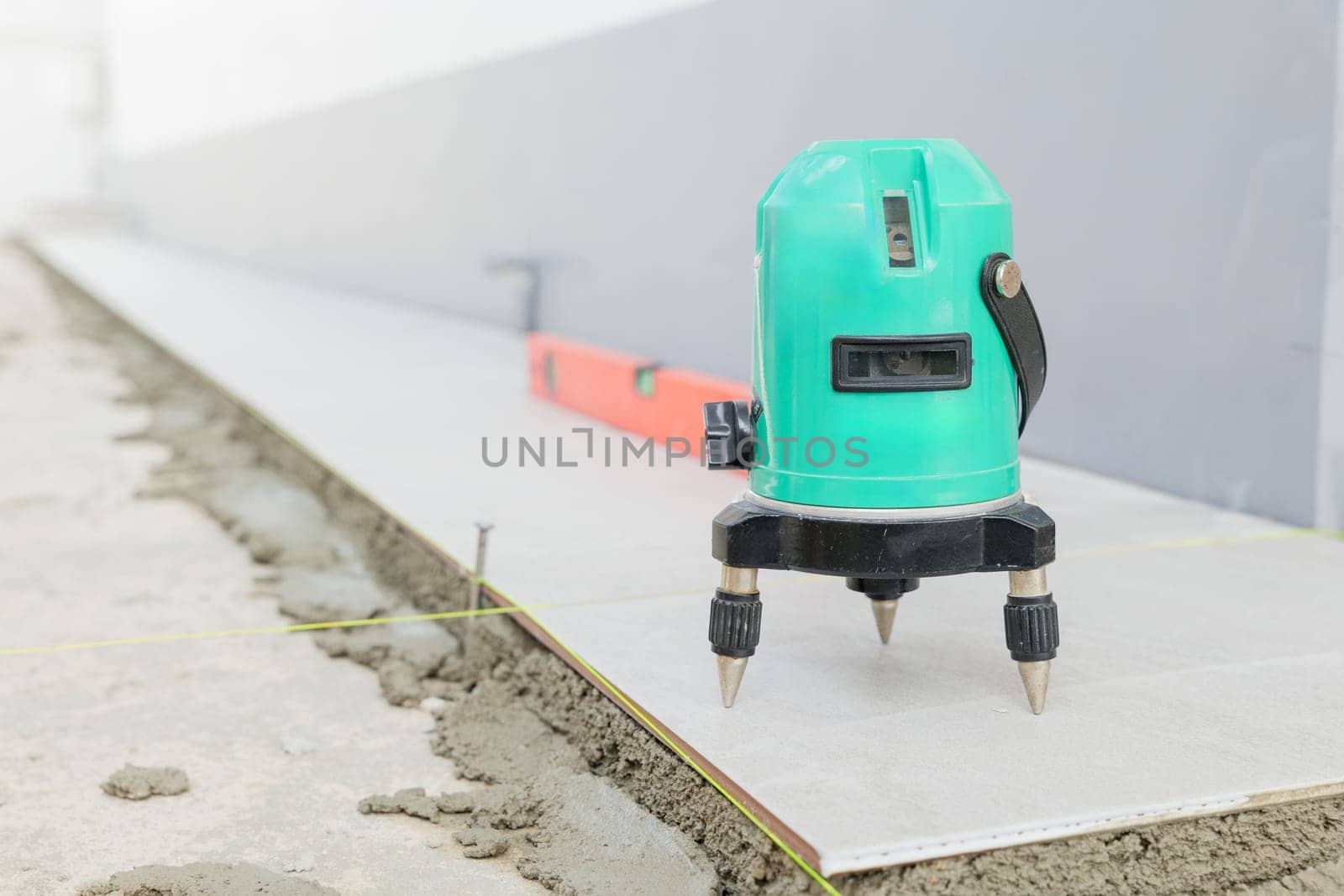Professional construction tools, green laser level positioned on a tripod for accurate ceramic tile placement on a white and gray floor.