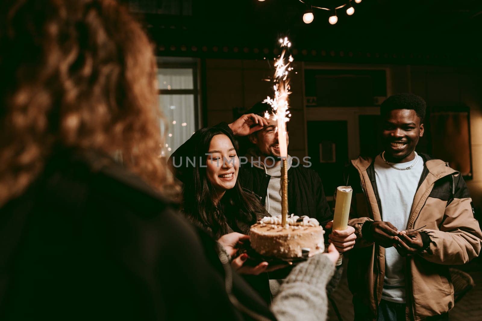 A group of friends gathers outdoors at night for a birthday celebration. One person presents a decorated cake with sparklers, while others smile and join in the festivities.