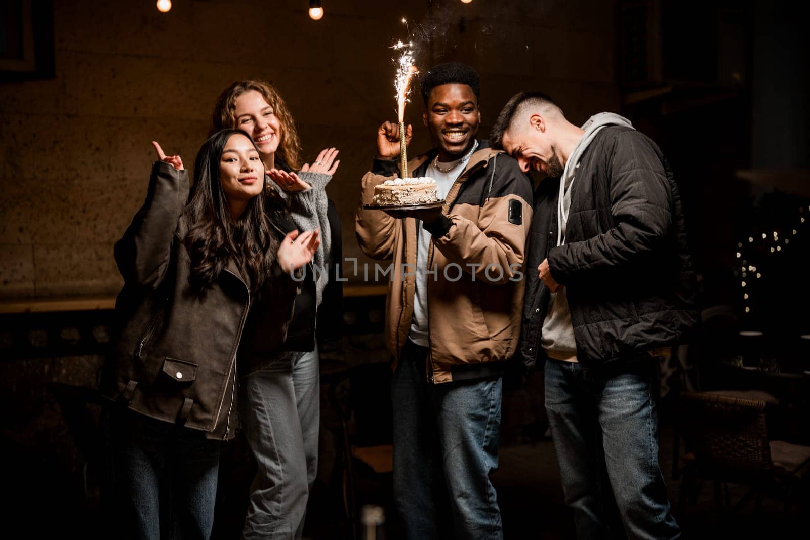 A group of friends enjoys a fun evening celebration outdoors, holding a cake with sparklers. Laughter and happiness fill the air as they share this special moment.
