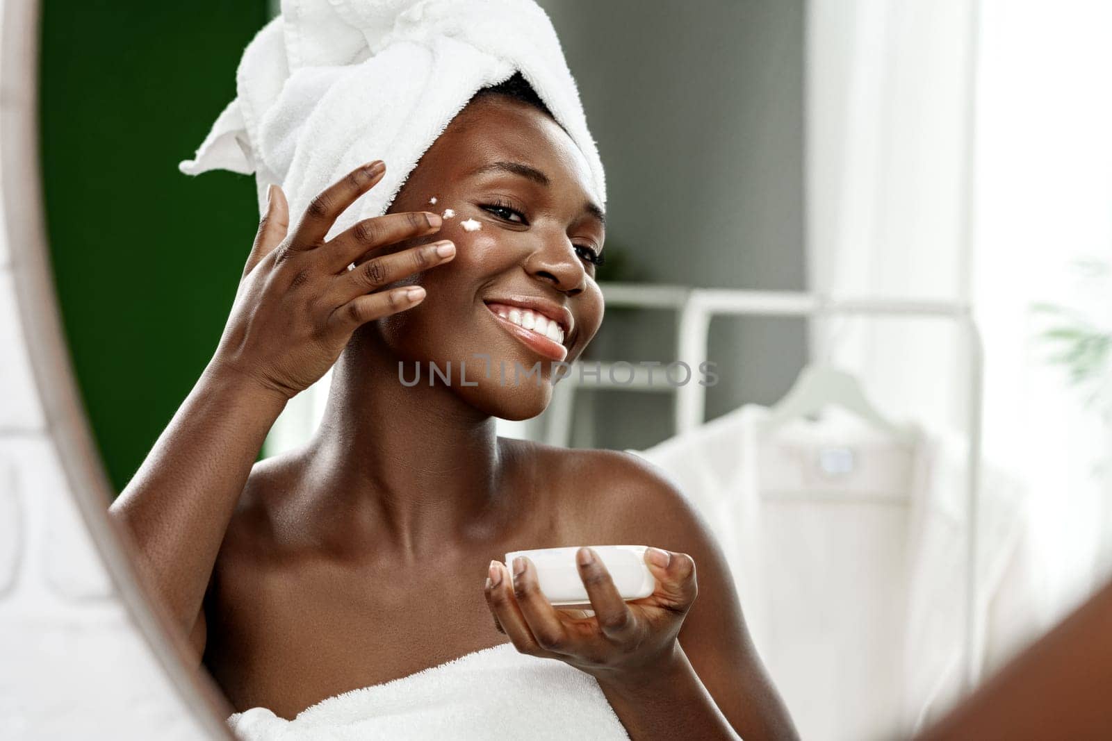 A young woman with a towel on her head smiles while applying cream to her face in a modern bathroom. Natural light highlights her cheerful morning routine.