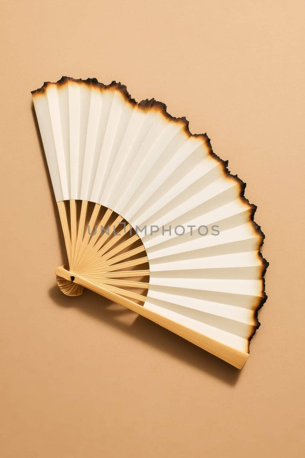 White paper fan with burnt edges lies on a neutral beige surface. A conceptual and symbolic image illustrating overheating, global warming, or fragile cooling efforts. Editorial or creative use